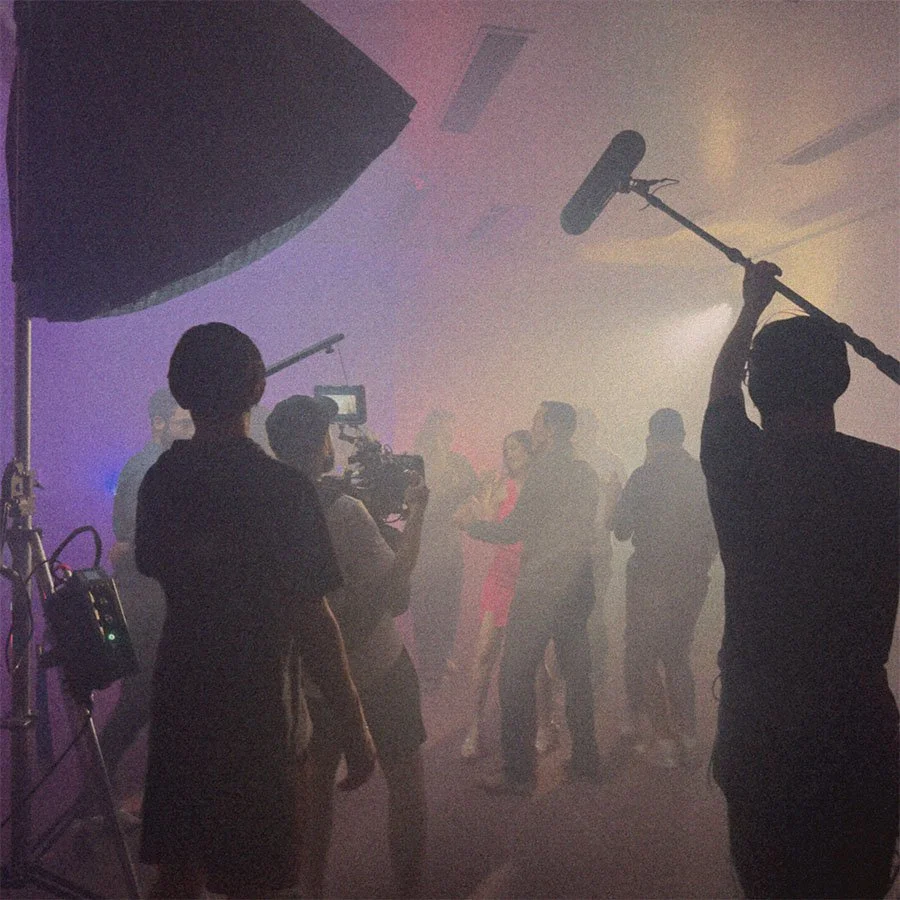Equipe de filmagem grava uma festa com pessoas dançando e se divertindo, com iluminação de estúdio e fumaça no ambiente.