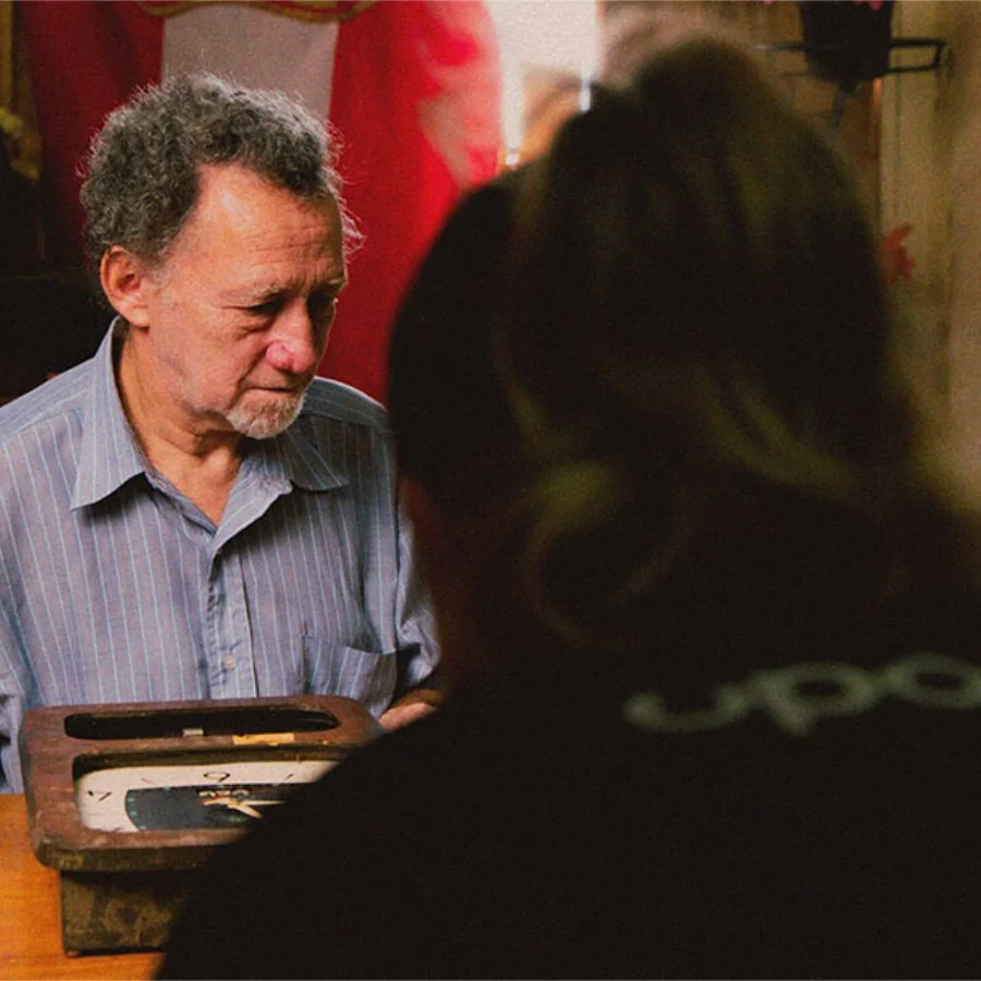 Homem idoso com cabelo grisalho, usando camisa azul, olhando para um tabuleiro de jogo, com uma mulher de cabelo escuro na frente, em um ambiente interno.