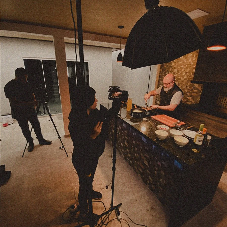 Cena de gravação de vídeo na cozinha, com uma pessoa segurando uma câmera e uma pessoa cozinheiro preparando comida sob uma grande sombrinha de iluminação, em um ambiente interno com duas pessoas ao fundo.