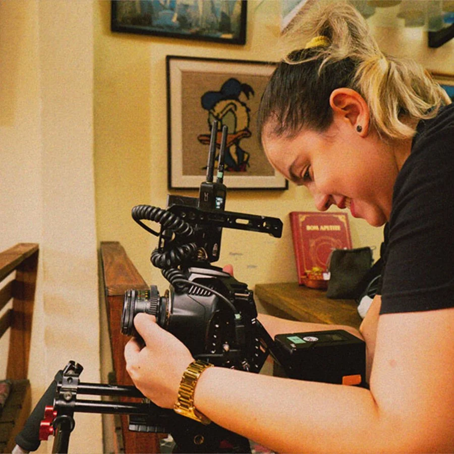 Pessoa ajustando uma câmera de vídeo com um medidor de luz em uma mesa, em um ambiente com quadros na parede.