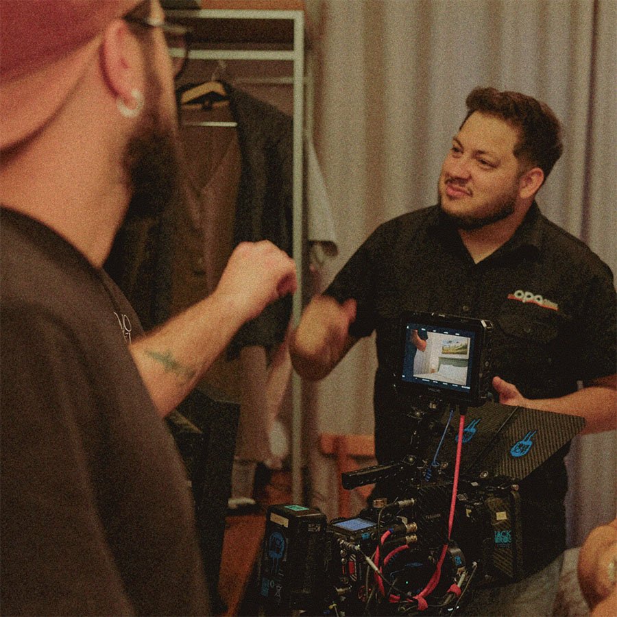 Homem com camiseta preta e laptop em mãos conversando com pessoa com cabelo ruivo e óculos em câmera de filmagem.