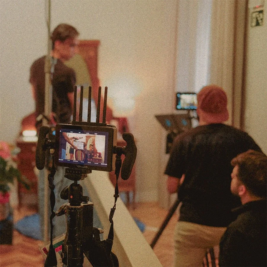 Equipe de gravação de um vídeo ou making-of com duas câmeras focadas em uma pessoa em um cenário interno.