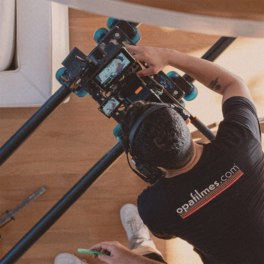 Pessoa filmando com uma câmera de cinema de alta qualidade, usando fones de ouvido enquanto trabalha em um plano de filmagem, vestindo uma camiseta preta com o site opafilmes.com, em um ambiente interno com piso de madeira.