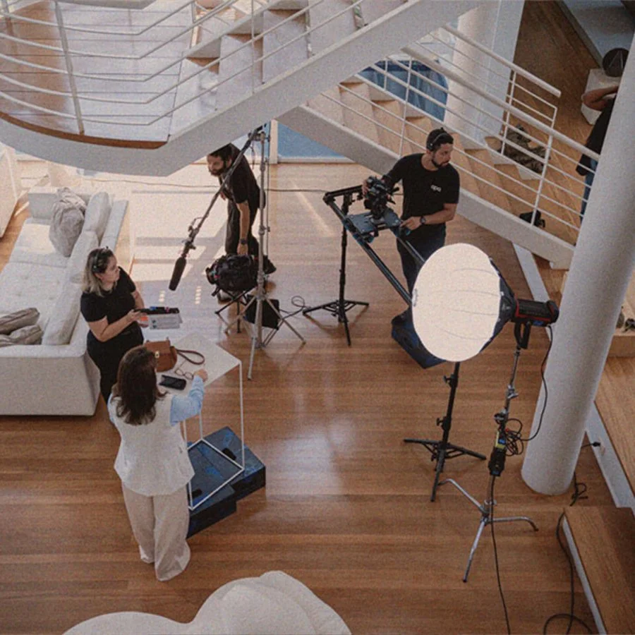 Equipe de filmagem em um estúdio com iluminação profissional, câmeras, tripés, e equipamentos de gravação, com uma mulher em pé ao centro e outras pessoas operando os equipamentos.