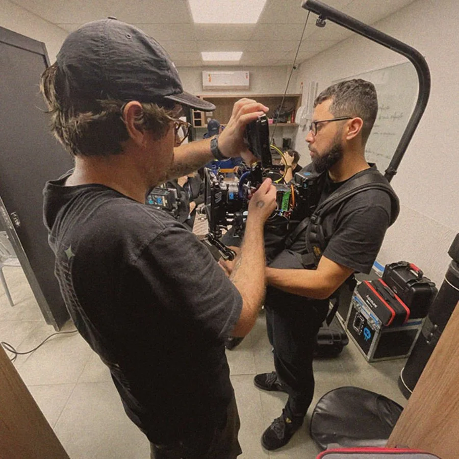 Duas pessoas trabalhando na preparação de uma câmera de cinema em um ambiente interno, com equipamentos ao redor.