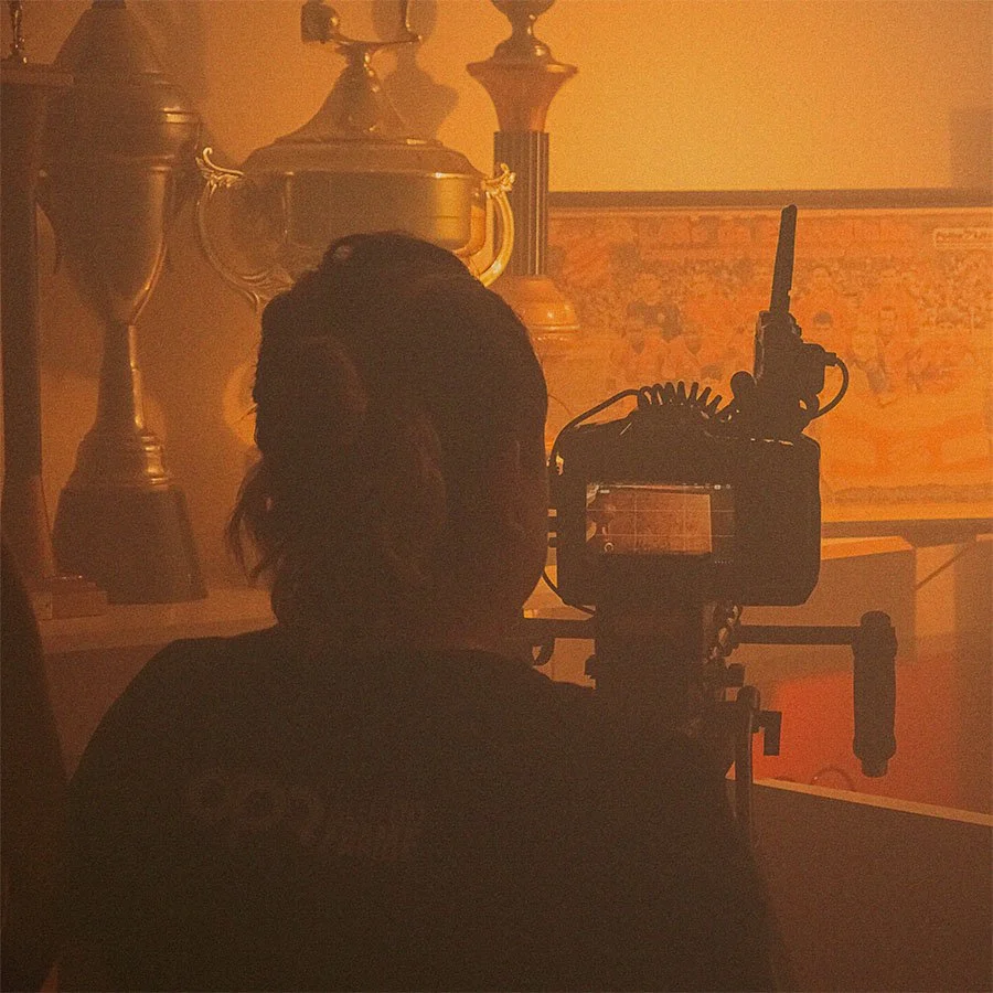 Pessoa operando uma câmera de filmagem em um ambiente com iluminação quente e objetos decorativos ao fundo.
