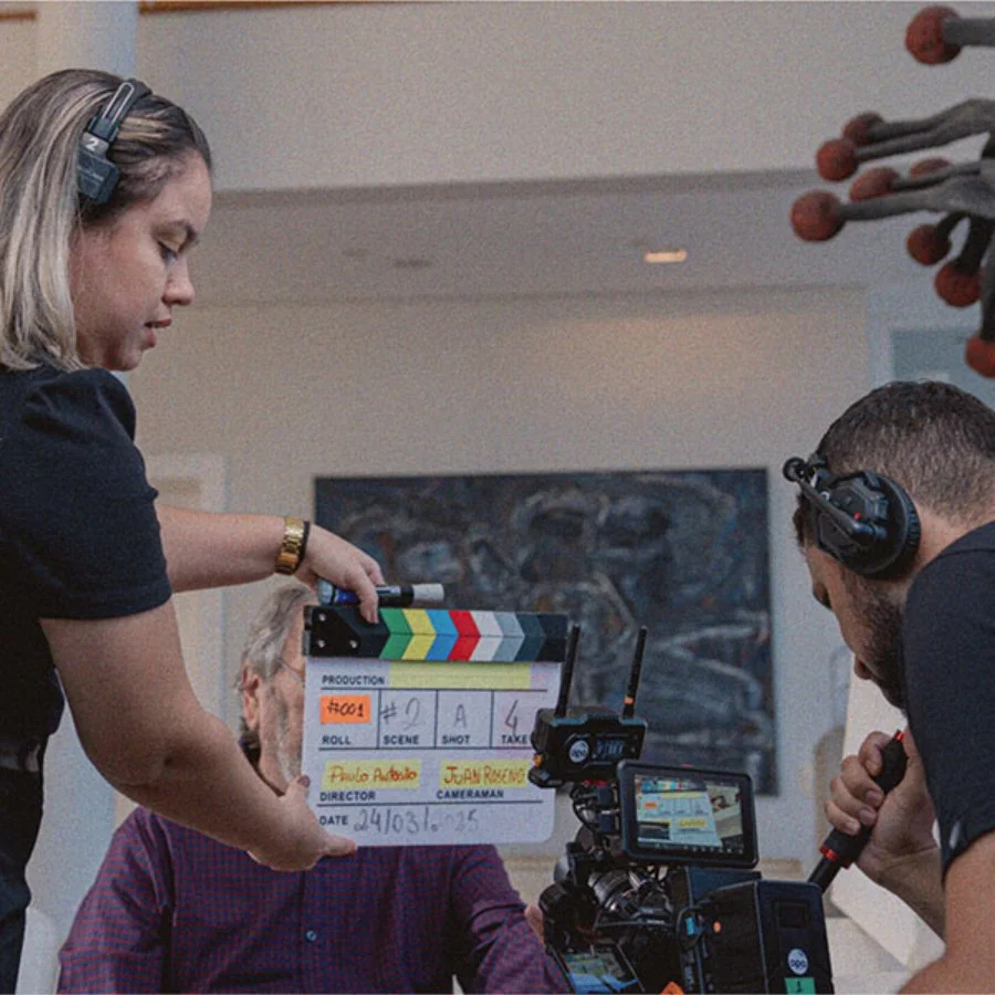Equipe de produção de filmagem em um set de filmagem, com uma cinegrafista segurando uma claquete e um cinegrafista operando uma câmera.