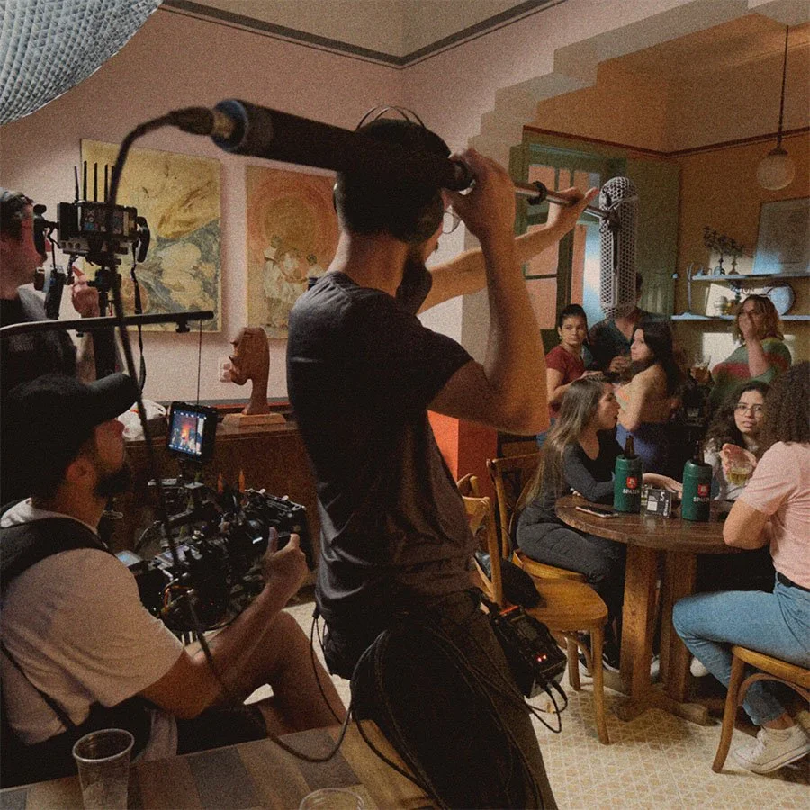 Pessoas filmando uma cena em um restaurante, com uma câmera de cinema e um microfone boom em destaque, enquanto outras pessoas conversam e interagem.