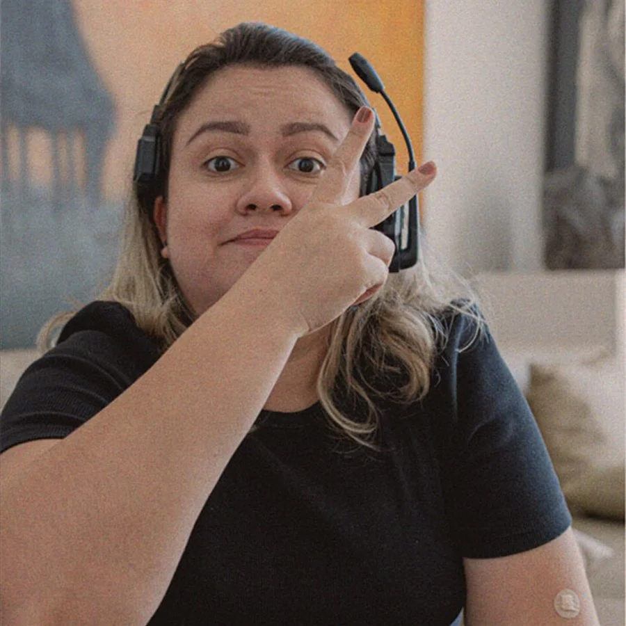 Mulher com cabelo castanho claro e cabelo solto, usando fone de ouvido com microfone, fazendo um gesto de paz com a mão, em um ambiente interno.