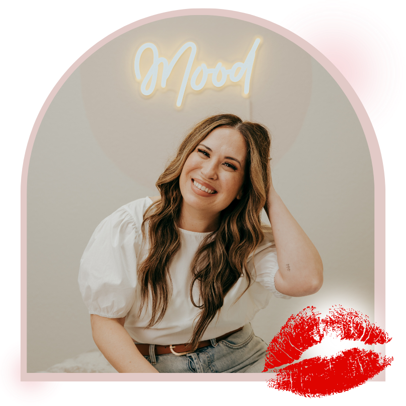 A smiling woman with long, wavy brown hair sitting indoors, touching her hair, with a neon sign that says 'Mood' behind her and a red lipstick kiss mark in the bottom right corner.