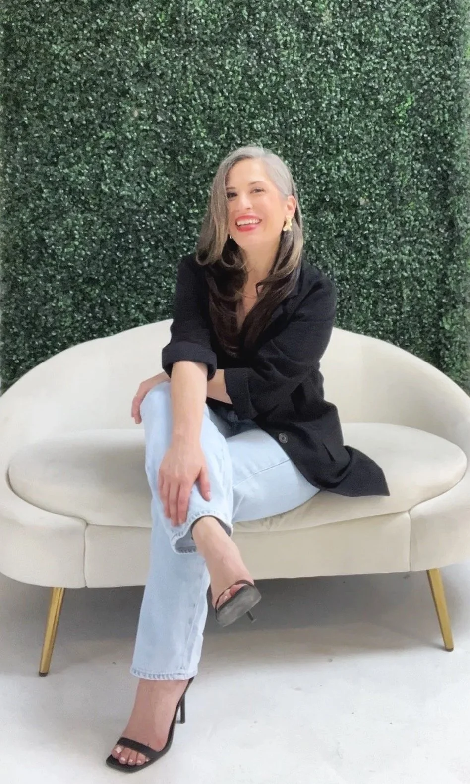 A woman with long brown hair,  sitting on a white couch with a green background wearing black blazer and jeans