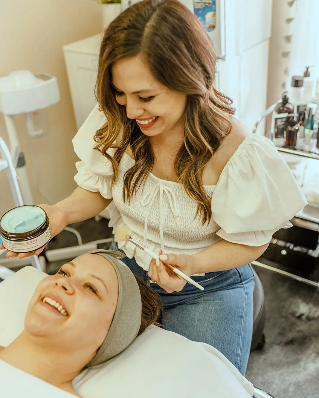 girl with brown hair holding a facial brush and blue mask to apply on woman