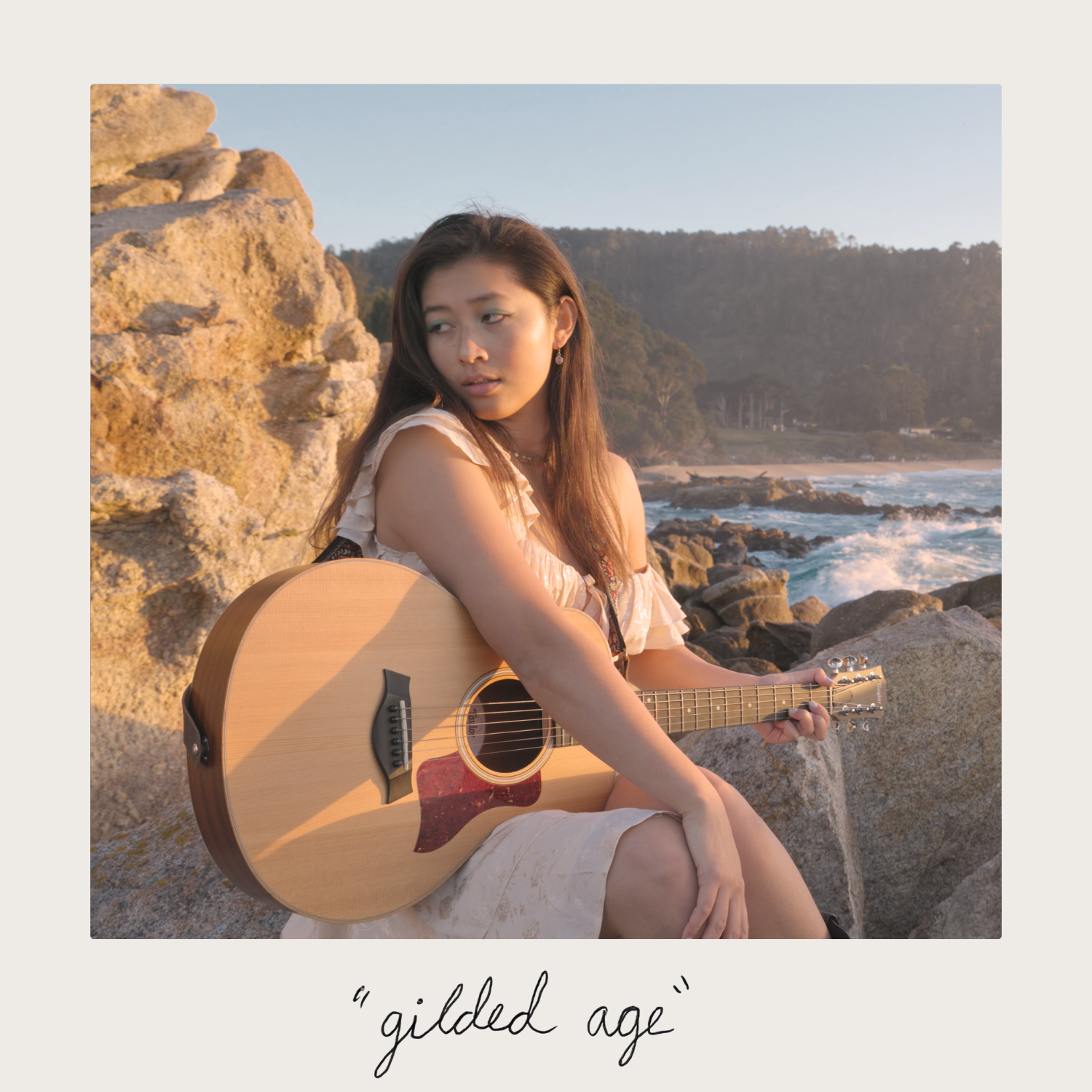 A young woman with long brown hair sitting on rocks by the beach, holding an acoustic guitar, with hills and the ocean in the background at sunset.