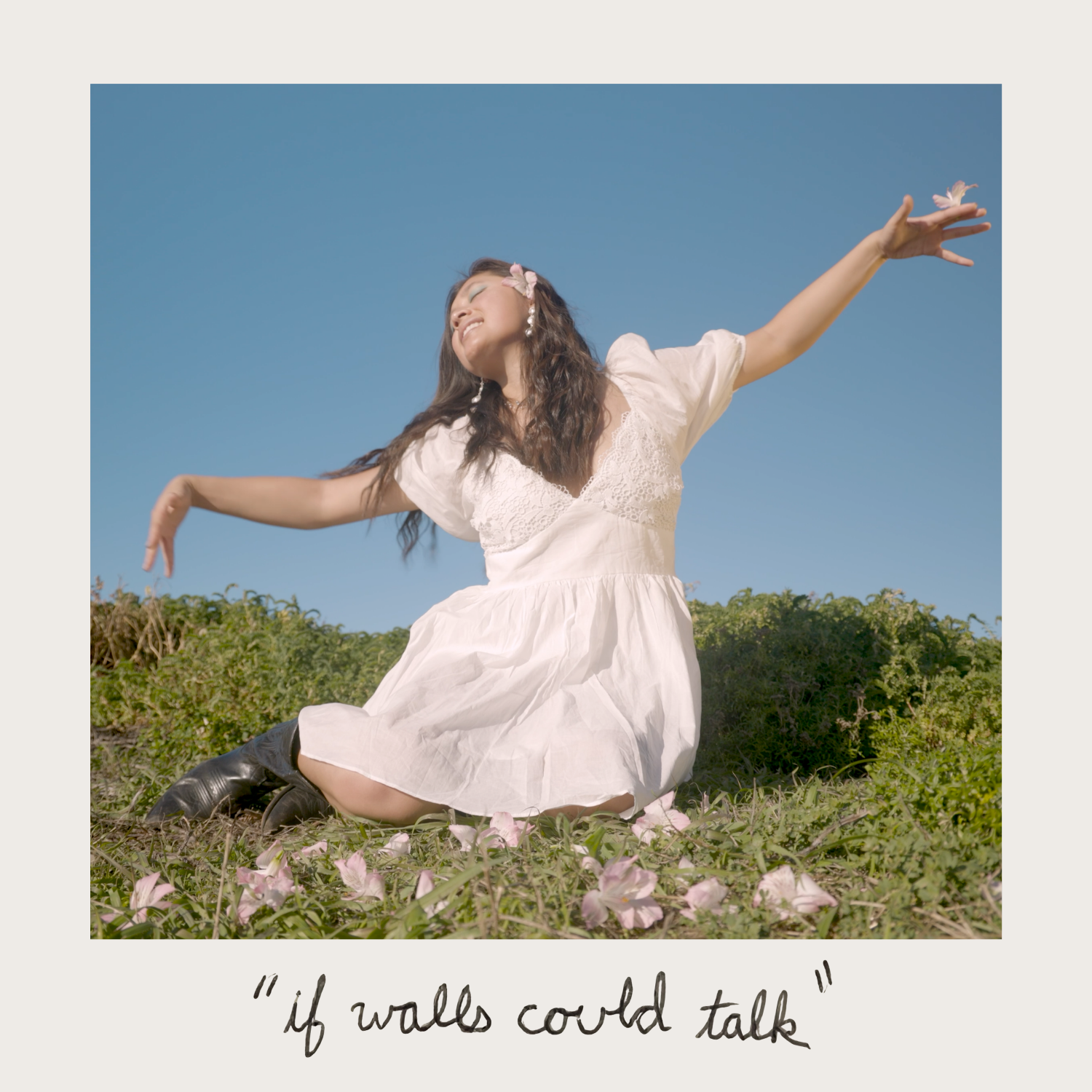 A young woman smiling, with arms outstretched, wearing a white dress and black boots, sitting on grass with pink flowers, under a blue sky, with the quote 'if walls could talk' written at the bottom.