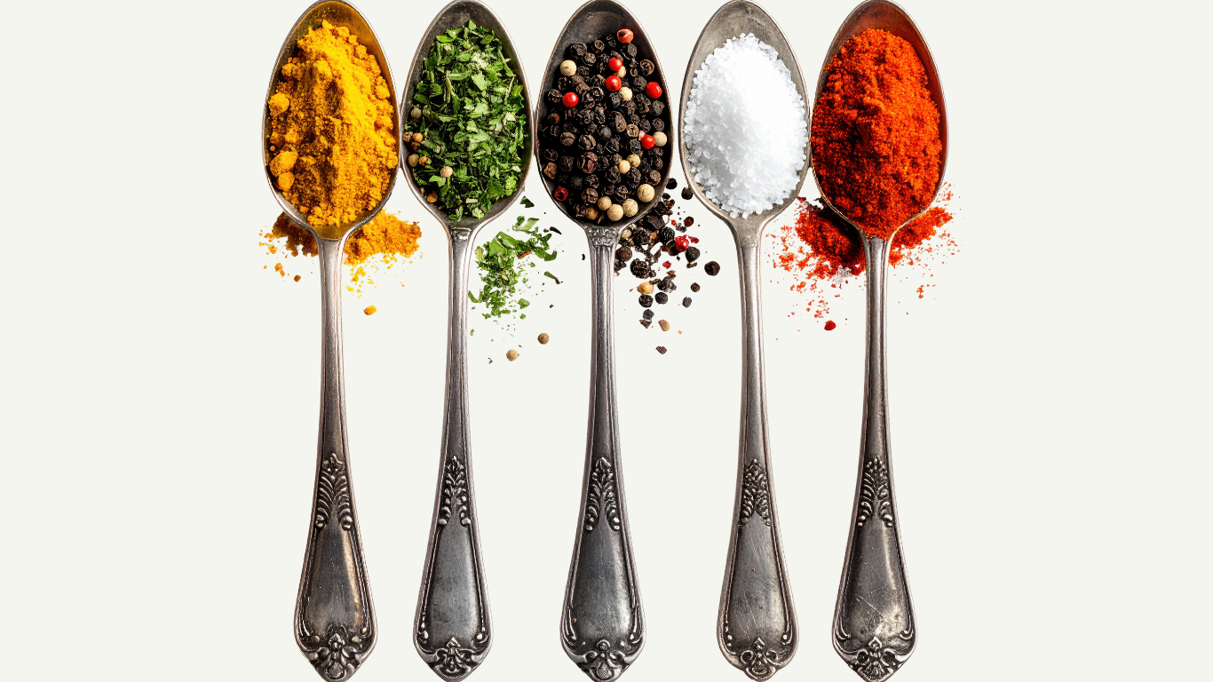 Five silver spoons filled with different spices and herbs, arranged in a row on a white background.