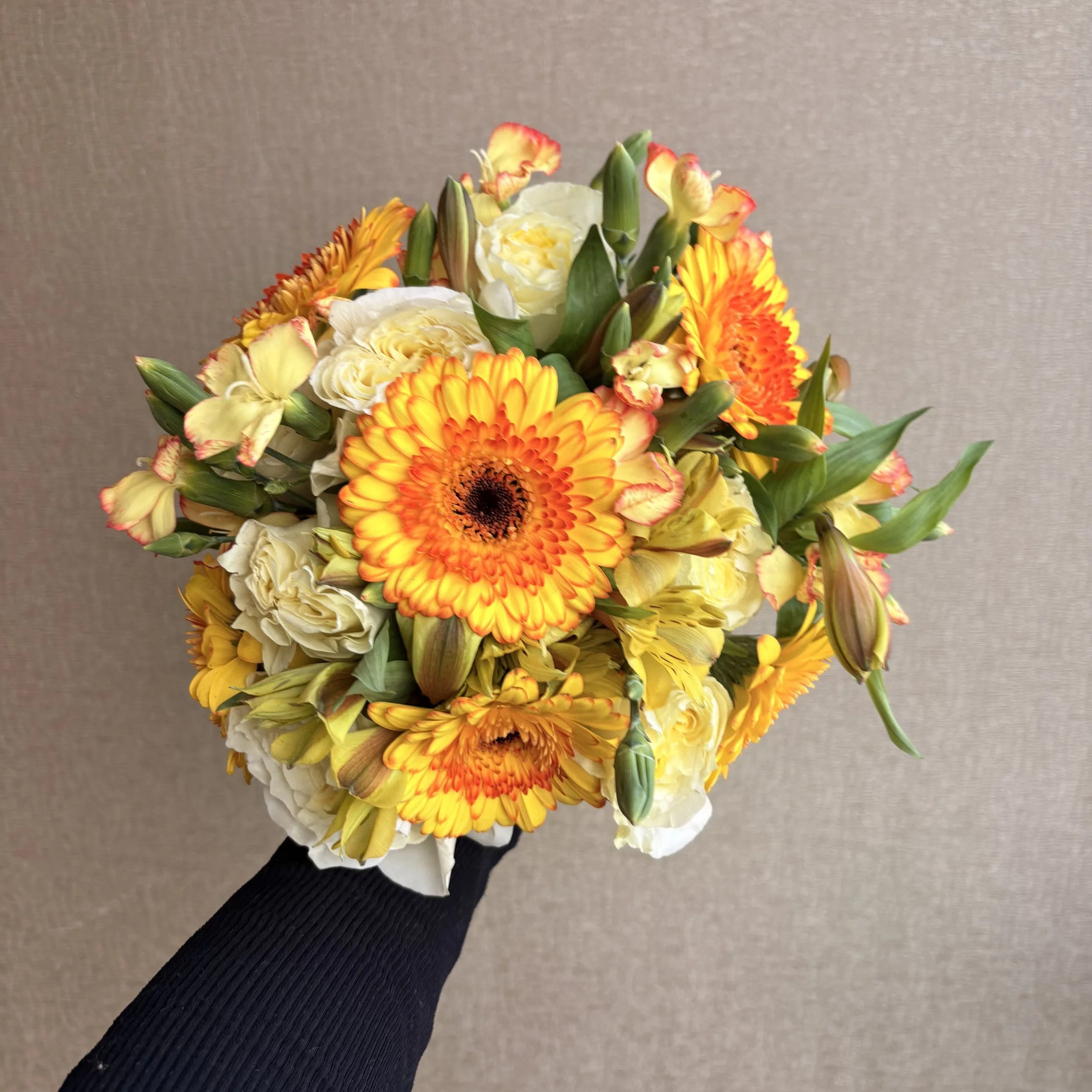 A hand in a black sleeve holding a bouquet of orange and yellow gerbera daisies, cream roses, and yellow lilies against a neutral beige background.