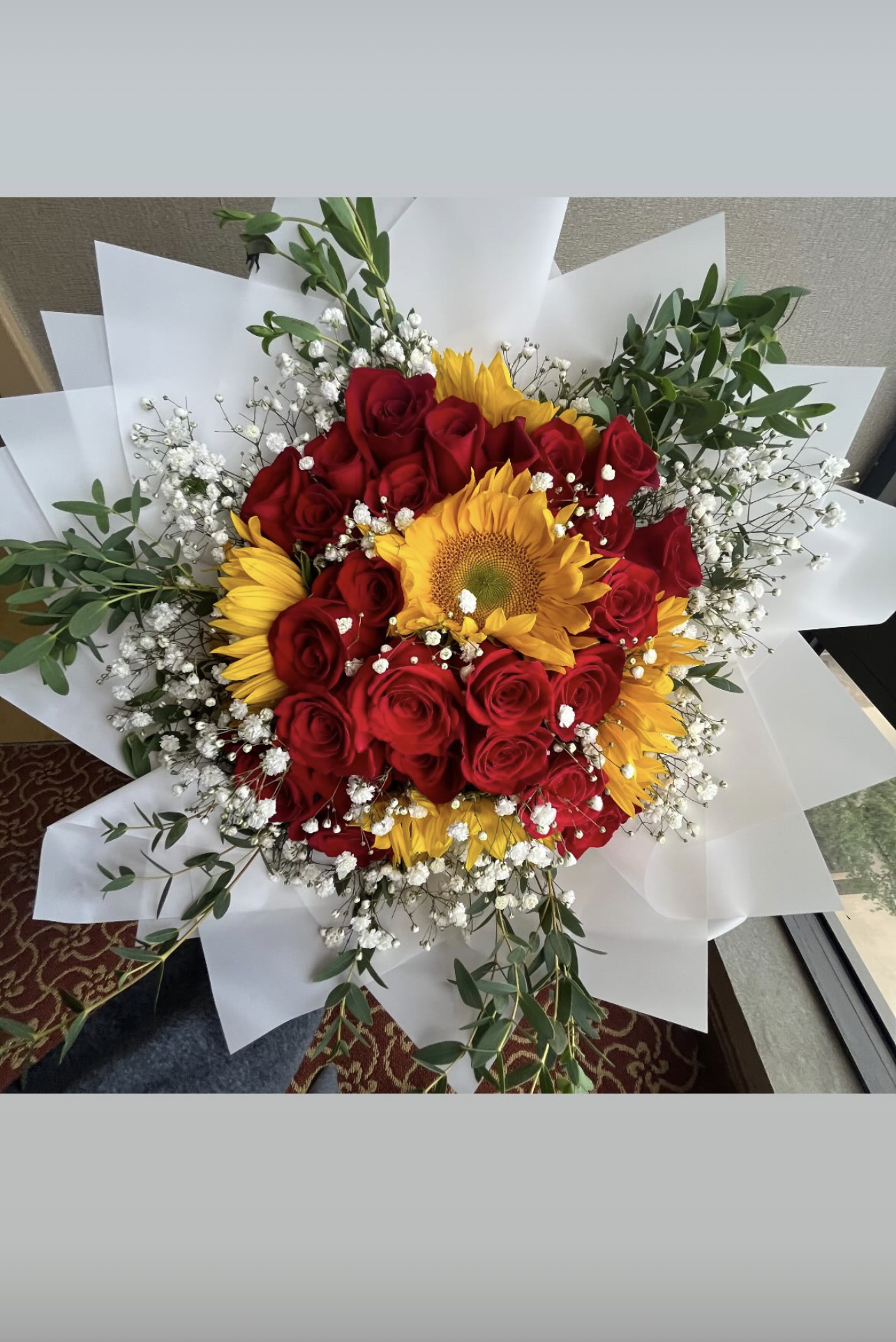 Bouquet of red roses, yellow sunflowers, white baby's breath, and green foliage wrapped in white paper.