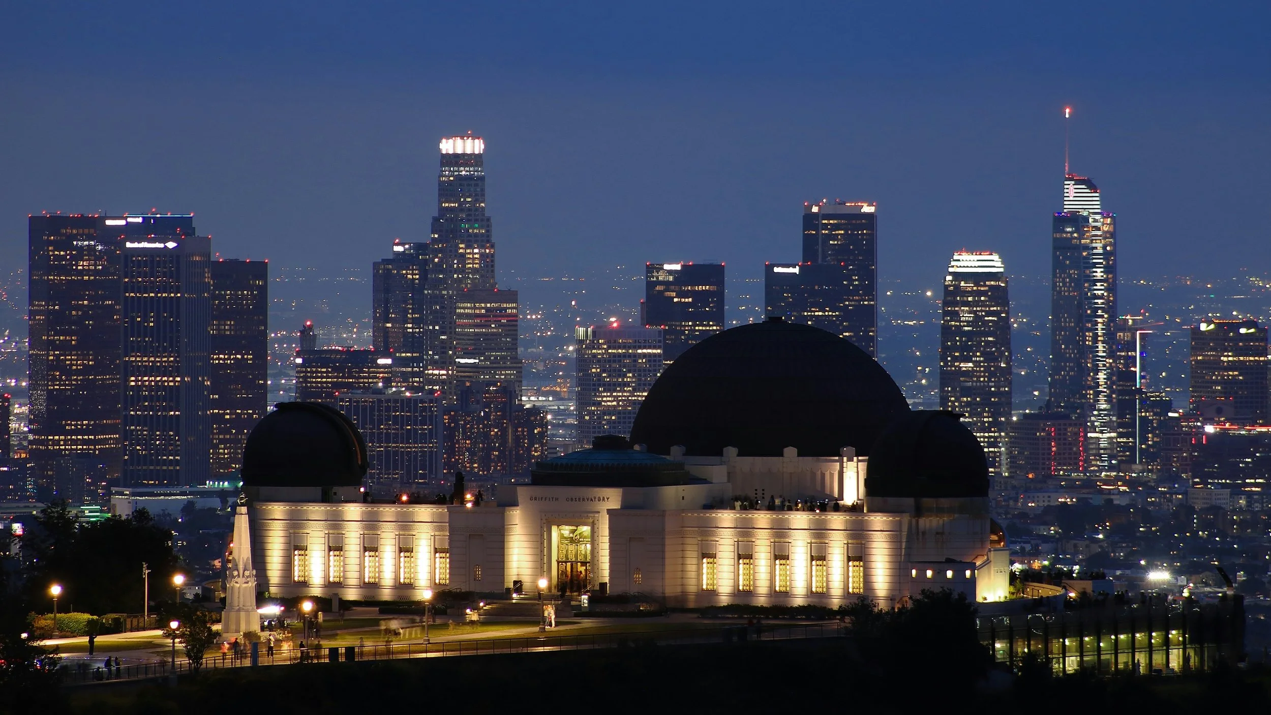 Nighttime view of the Griffith Observatory in Los Angeles with a city skyline of high-rise buildings illuminated in the background.