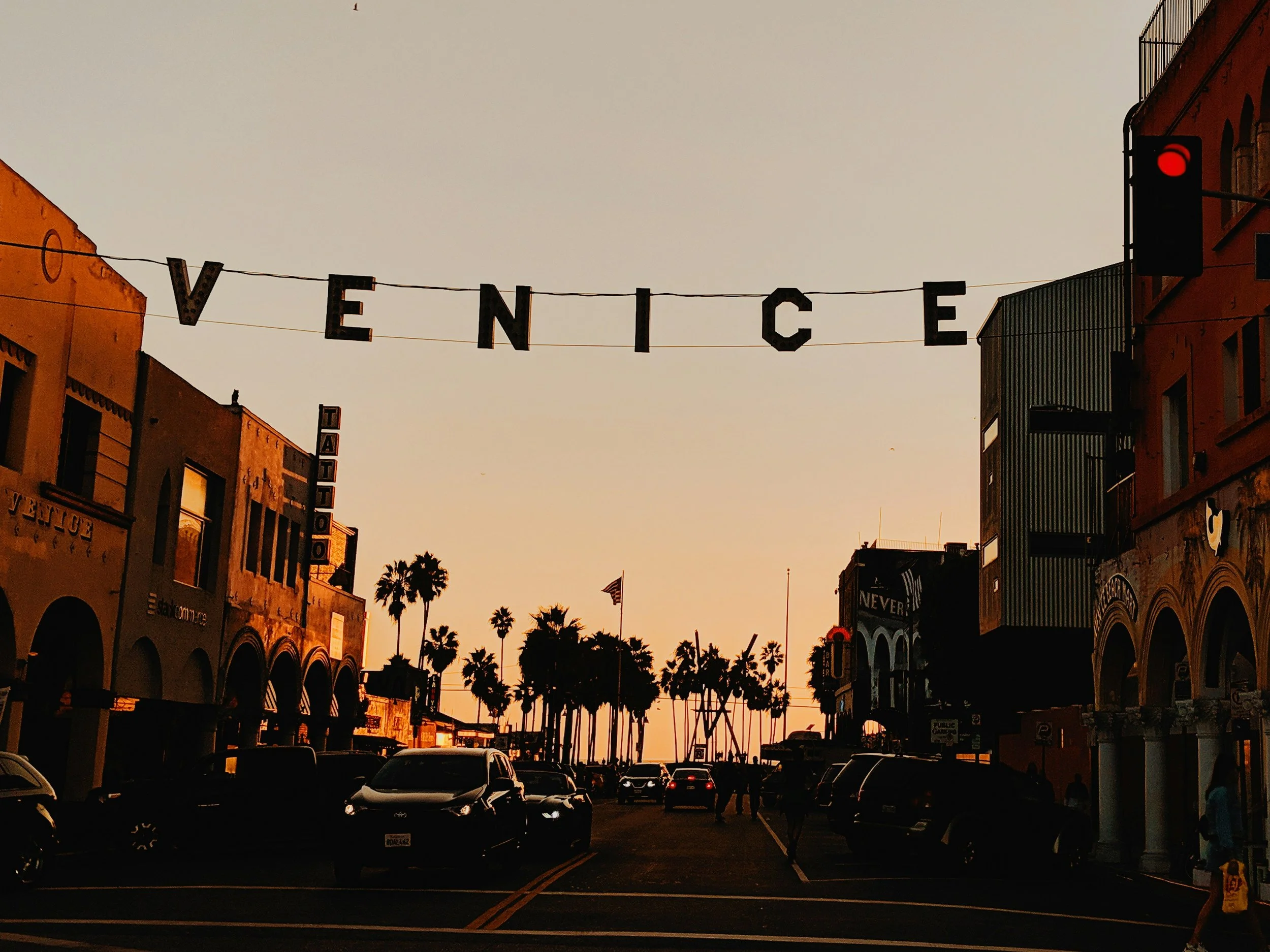 Street scene at sunset with palm trees, parked cars, colorful buildings, and a sign hanging across the street spelling 'Venice'.