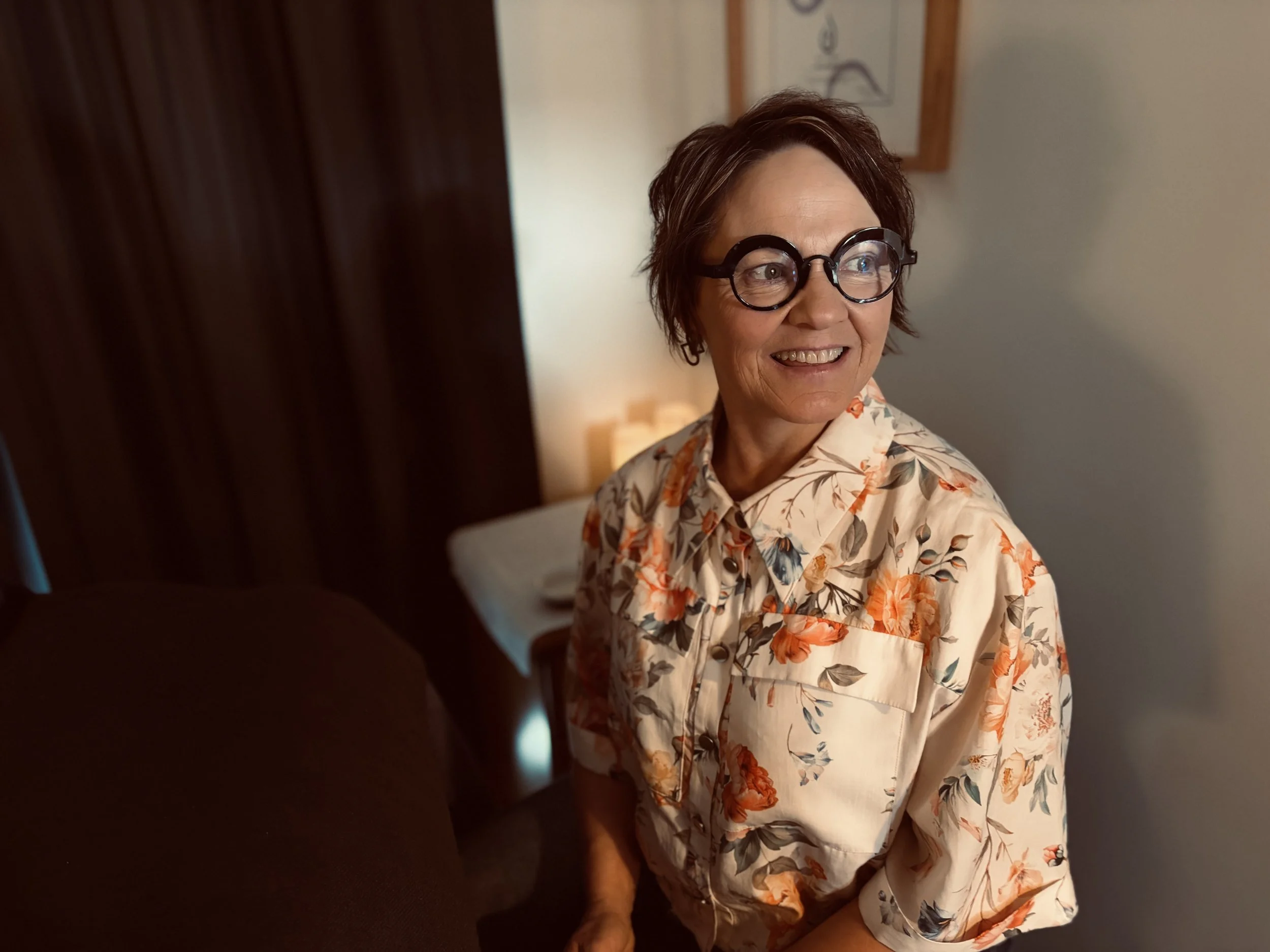 A woman with short brown hair, glasses, and a floral shirt smiling and looking to her right in a warmly lit room in a reflexology studio.