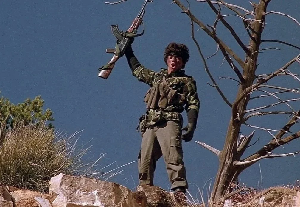 A young person standing on rocky terrain, holding a rifle overhead, wearing military-style clothing with a dark hoodie and gloves, next to a leafless tree against a cloudy sky.