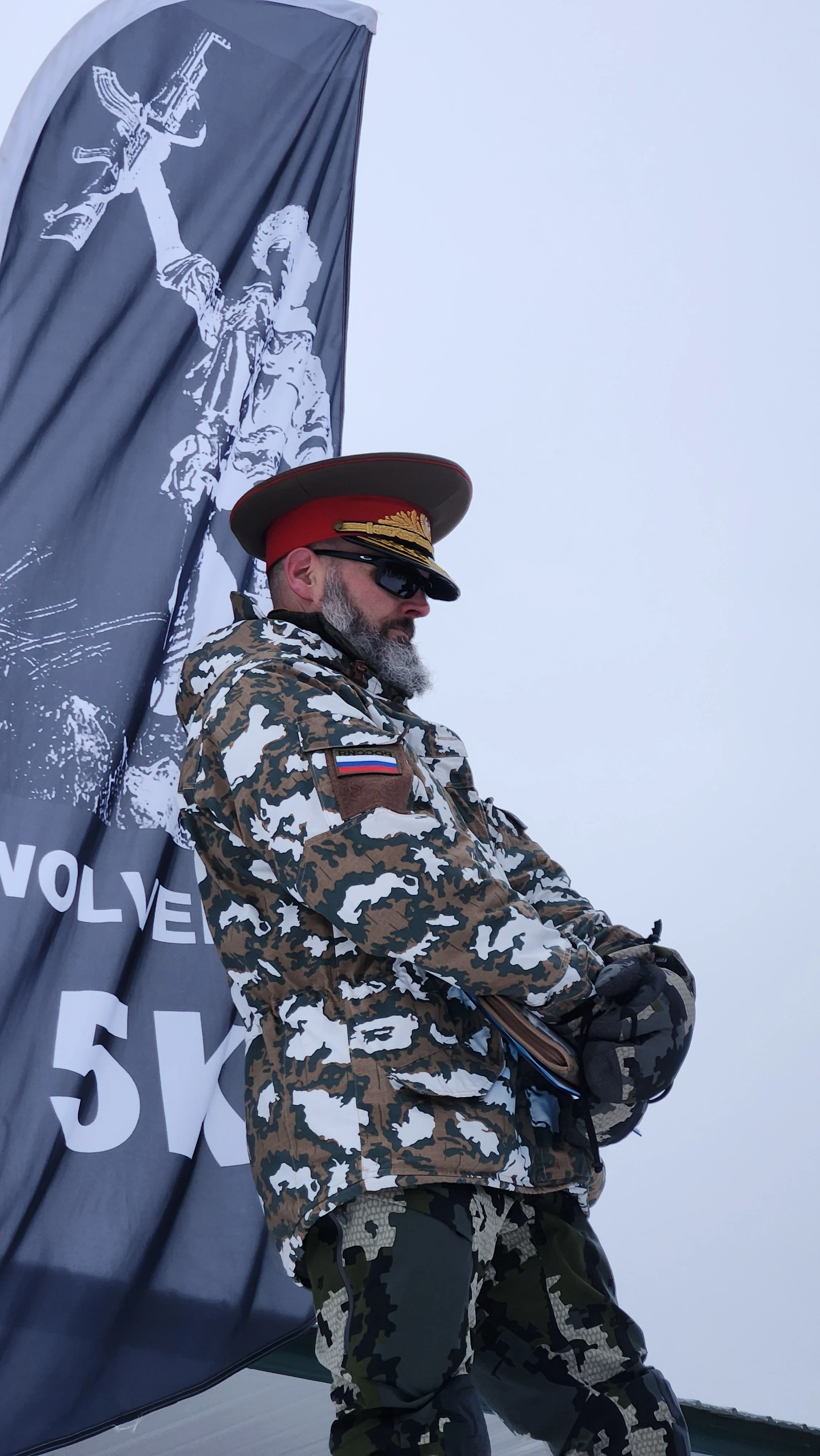 A man dressed in camouflage military attire standing next to a black banner with a white graphic of a soldier holding a gun and the words 'REVOLVE 5K'.