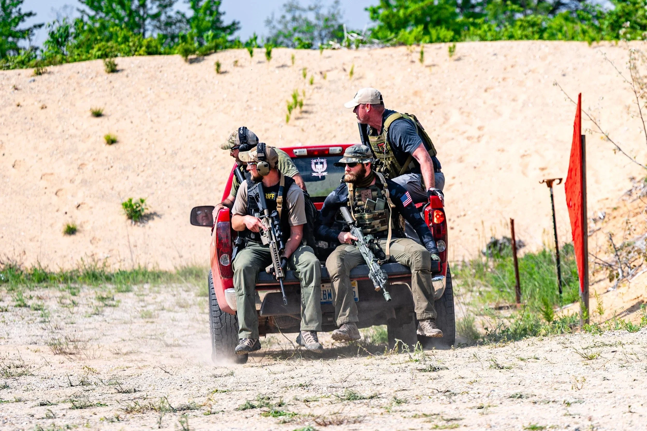 Four armed men in tactical gear sitting and standing in the back of a red pickup truck in a desert/field environment.