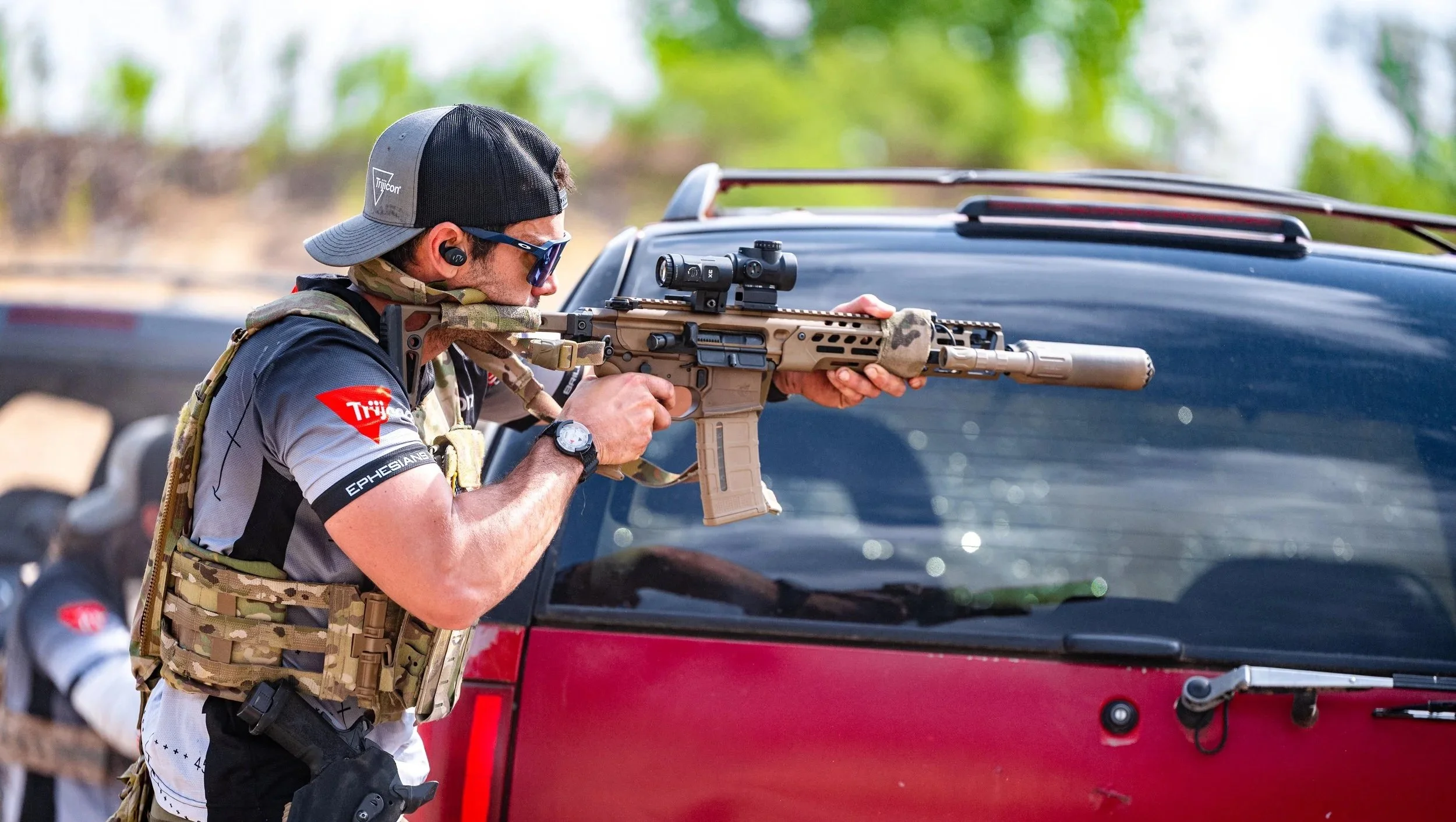 A man wearing a gray and black shirt, a camo tactical vest, a black baseball cap, sunglasses, and black ear protection, aiming a tan tactical rifle with a scope at a target, leaning against a black and red vehicle.