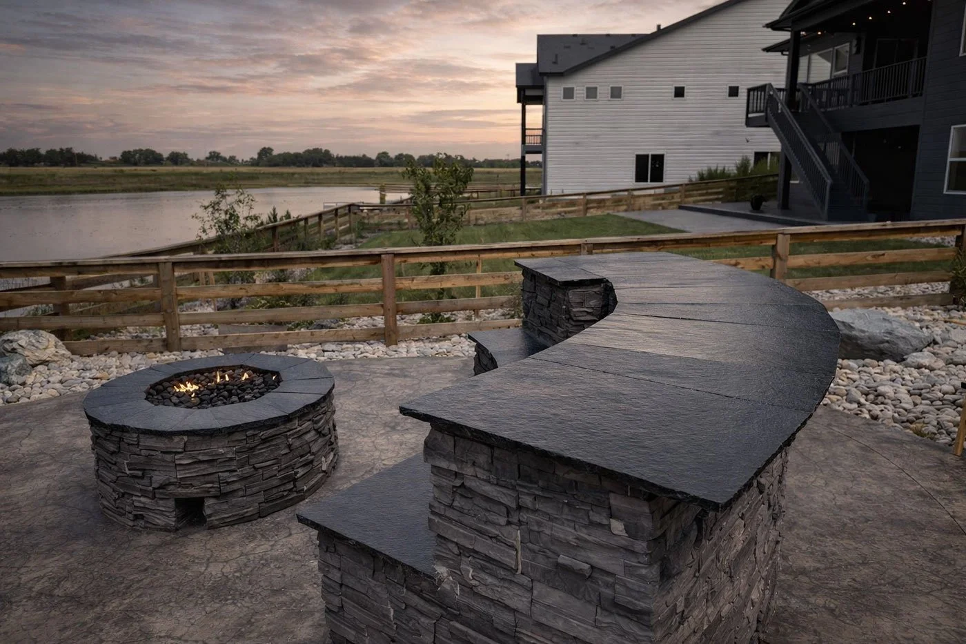 Outdoor patio with a curved stone fire pit and built-in seating, overlooking a river and houses at sunset.