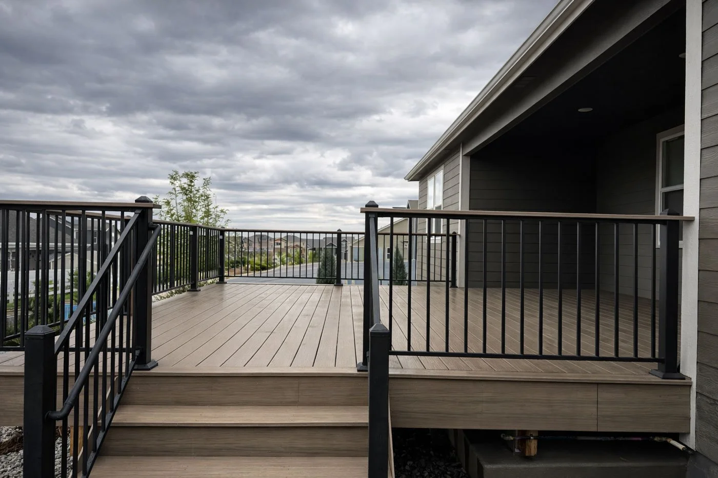A view of a residential deck with black metal railing and wood flooring, attached to a house with dark gray and white siding, overlooking a neighborhood street with cloudy skies.