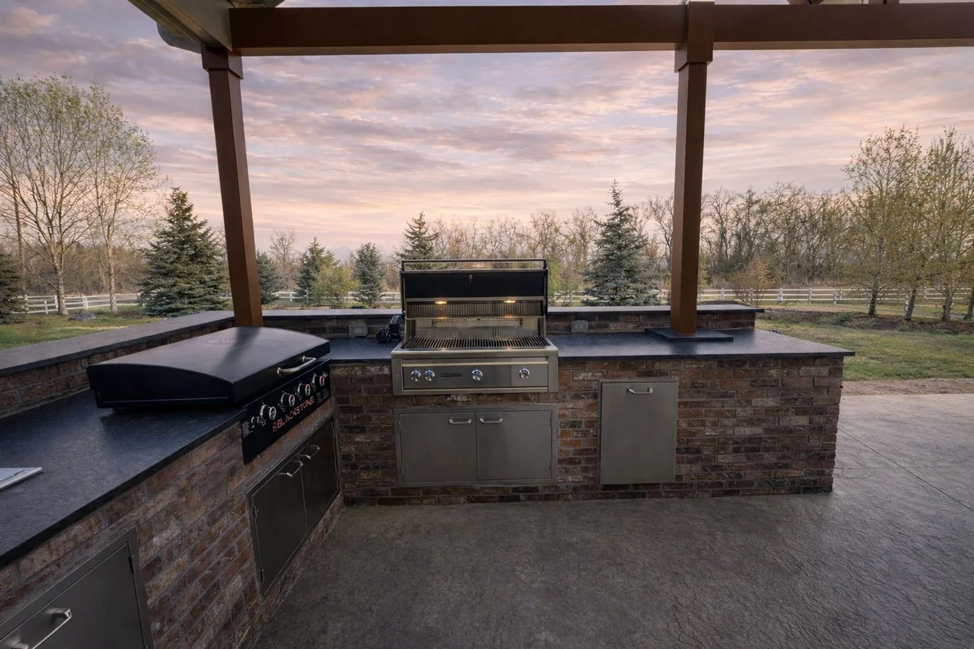 Outdoor kitchen with brick counter, stainless steel grill, black gas burner, and surrounding trees at sunset.
