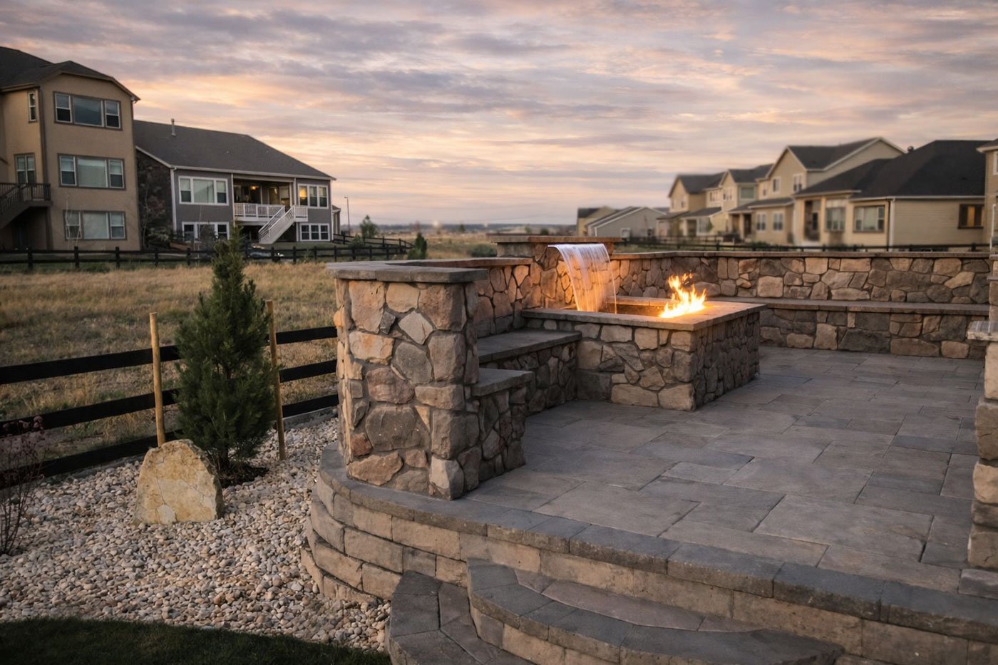 Backyard patio with stone fire pit and waterfall feature, surrounded by houses at dusk.