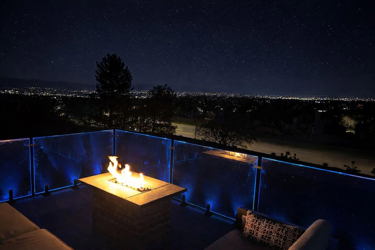 Nighttime view from a balcony with a lit fire feature, overlooking a city skyline under a starry sky.