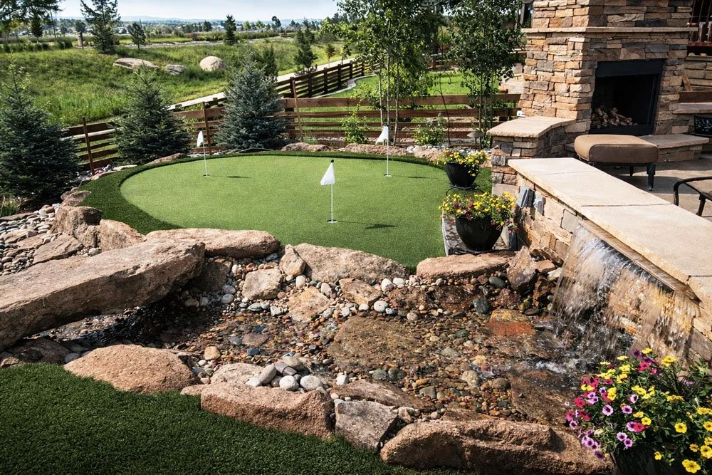 A backyard with a putting green, nearby rocks, a water feature with a waterfall, potted flowers, a stone fireplace, and outdoor seating.