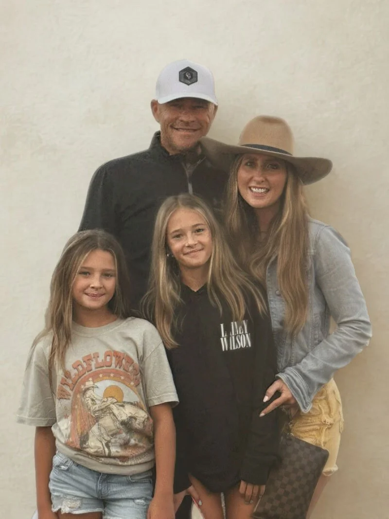A group of five people, two young girls and three adults, smiling and standing close together indoors against a plain wall. The adults are a man wearing a white cap and a woman with a large floppy hat, while the children are dressed casually.