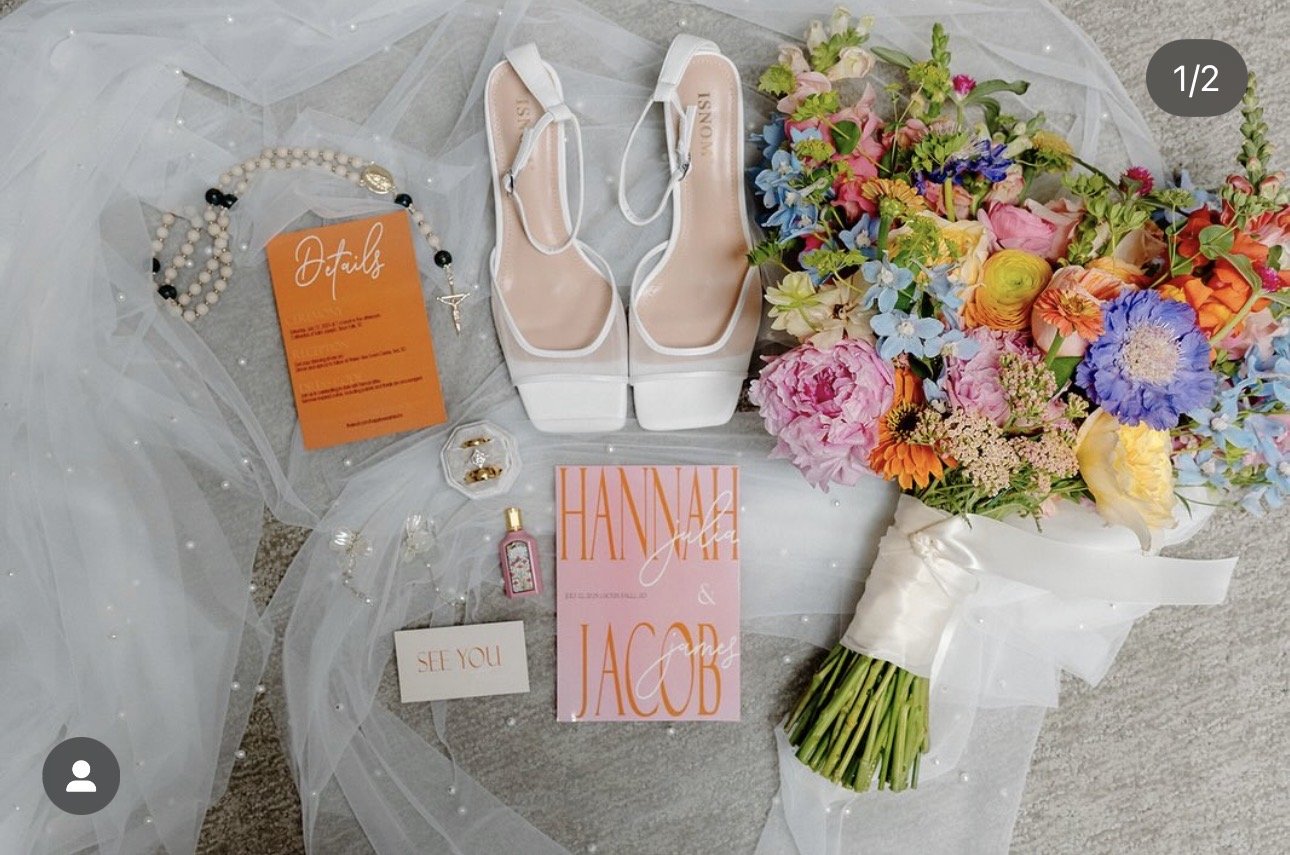 A wedding flat lay showing a bouquet of colorful flowers, white high-heeled shoes, jewelry, a pink invitation card with 'Hannah & Jacob,' a small perfume or oil bottle, a 'Details' card, a 'See You' note, and a sheer fabric with small pearl embellish