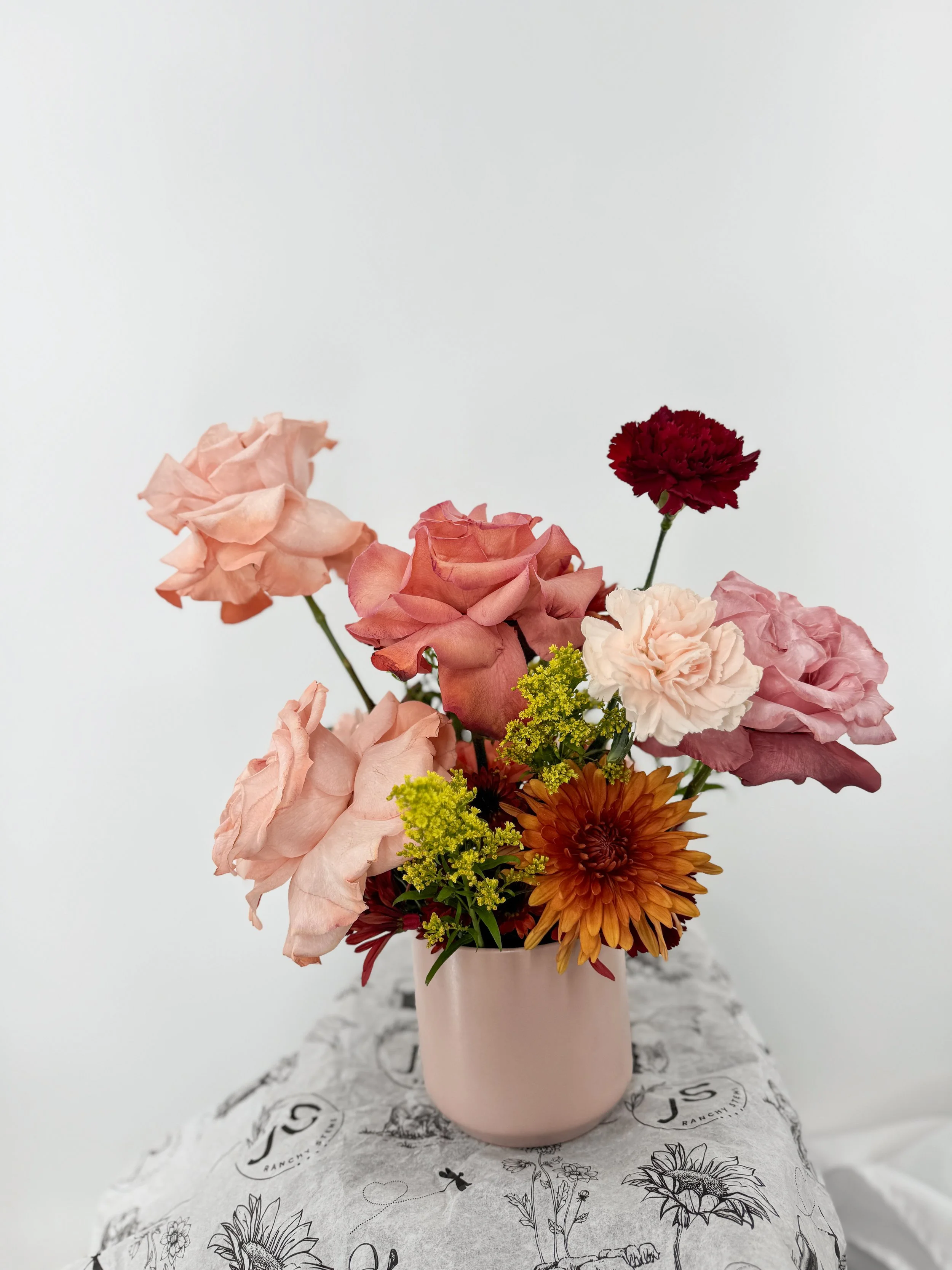 A pink ceramic vase with a mixed arrangement of pink, red, orange, and yellow flowers on a table with a floral-patterned cloth against a plain white background.