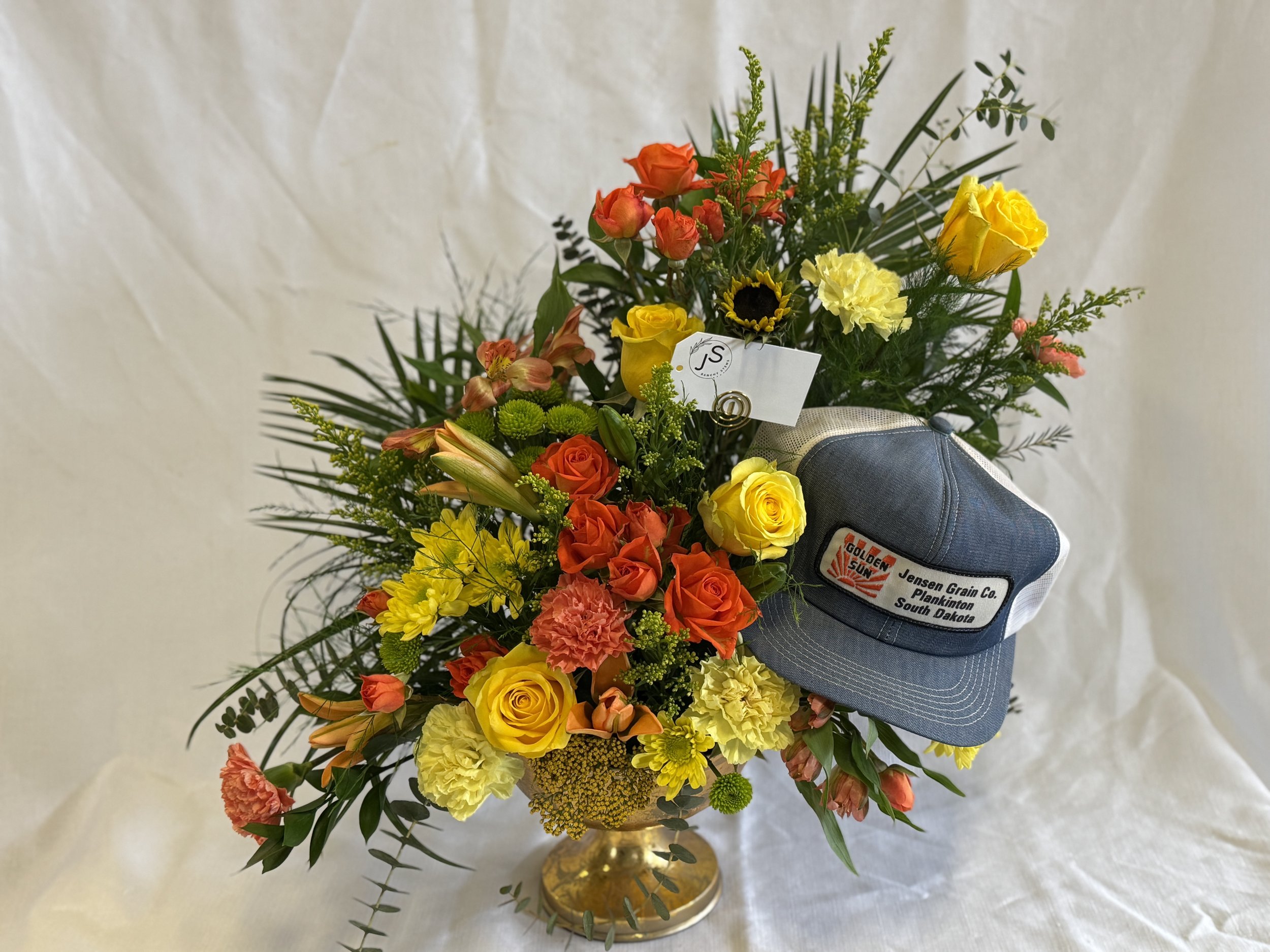 Colorful flower arrangement in a gold vase with yellow, orange, and pink roses, lilies, carnations, sunflowers, and various greenery, with a blue cap that has a patch reading "Jensen Grain Co. Plankinton South Dakota."