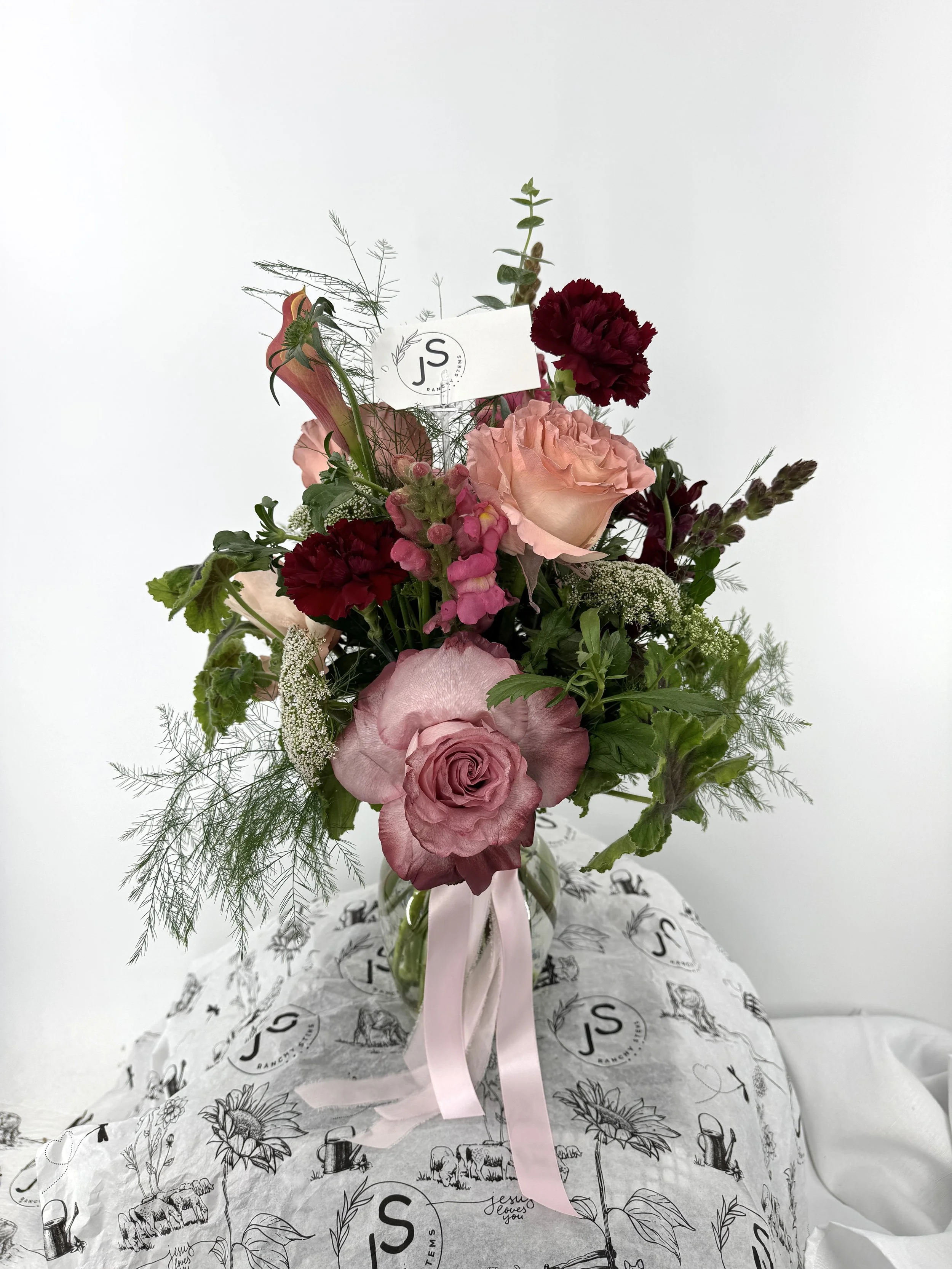 A bouquet of pink, red, and peach flowers with greenery in a glass vase, tied with a pink ribbon, placed on decorative paper with logo 'JS' and illustrations, against a plain white background.