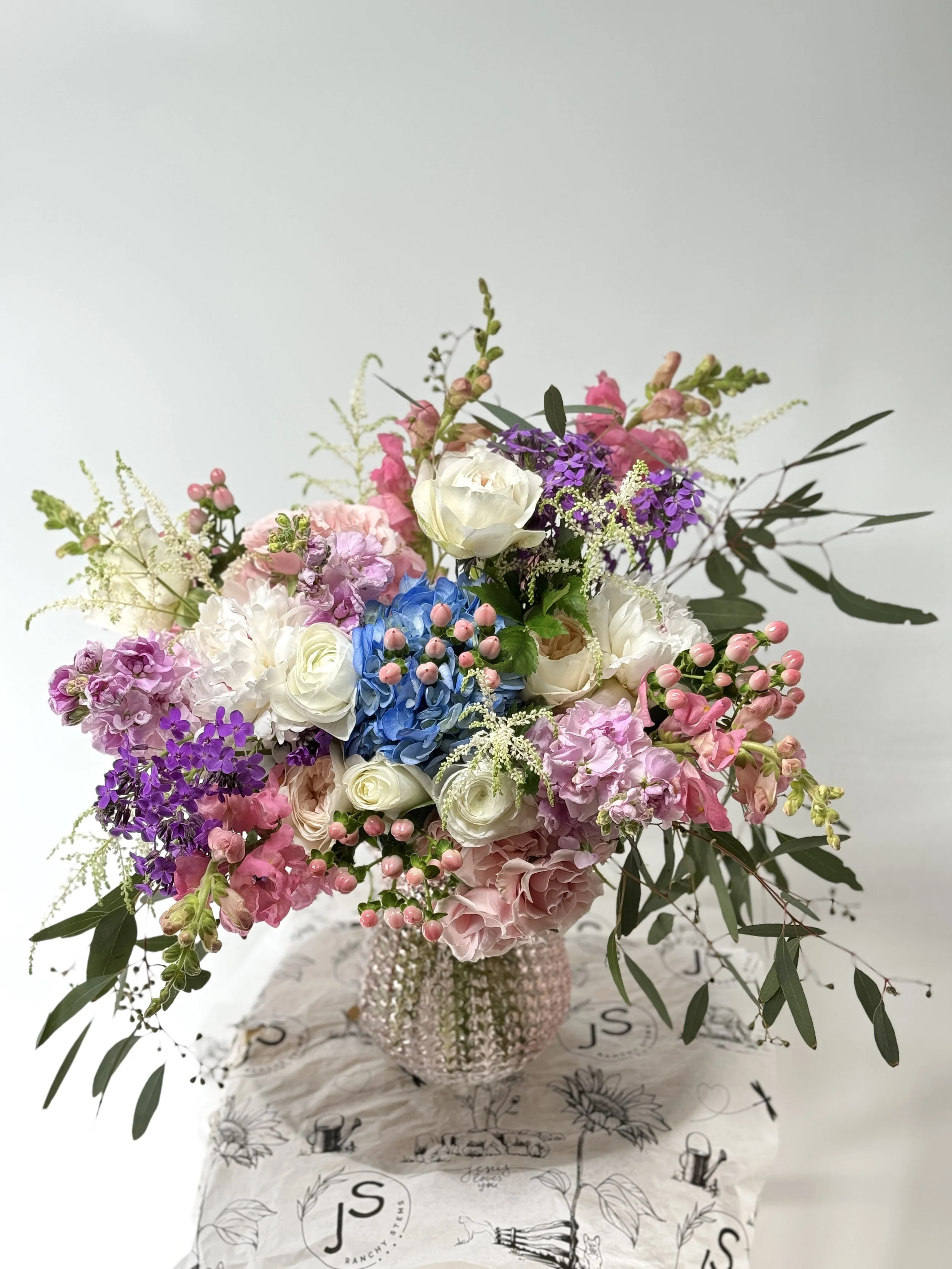 Beautiful floral arrangement with pink, white, purple, and blue flowers in a pink textured vase, placed on a white cloth with black and white sketches, against a plain white background.