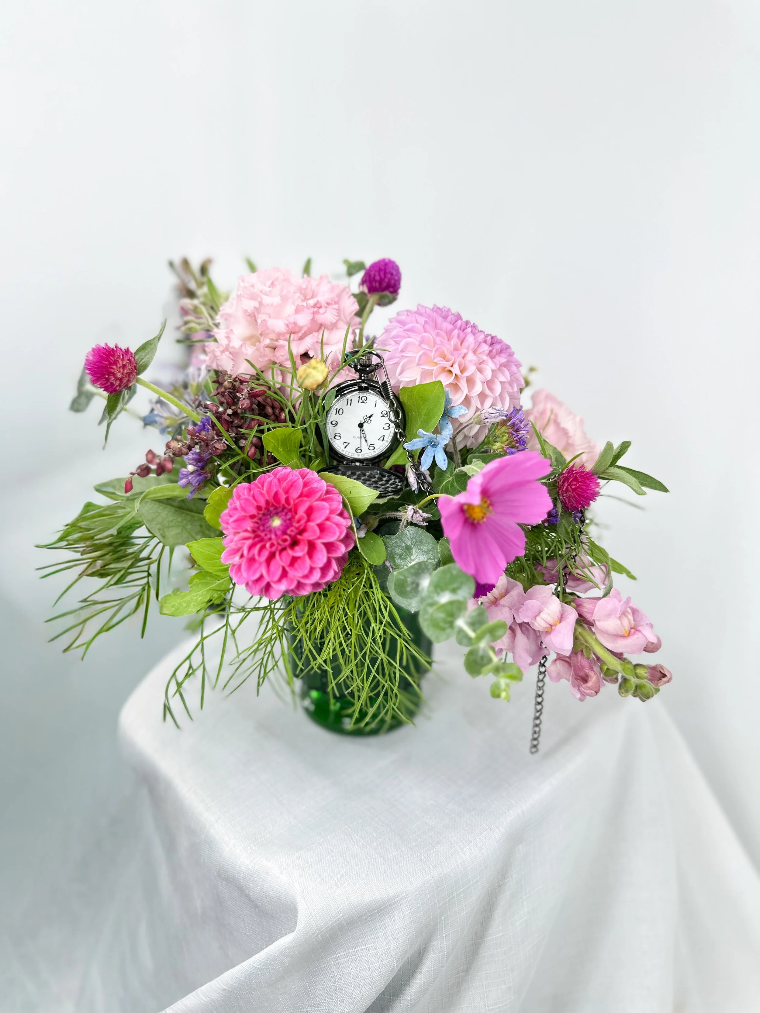 Flower arrangement in a green vase with a clock among pink, purple, and blue flowers on a white table with a white background.