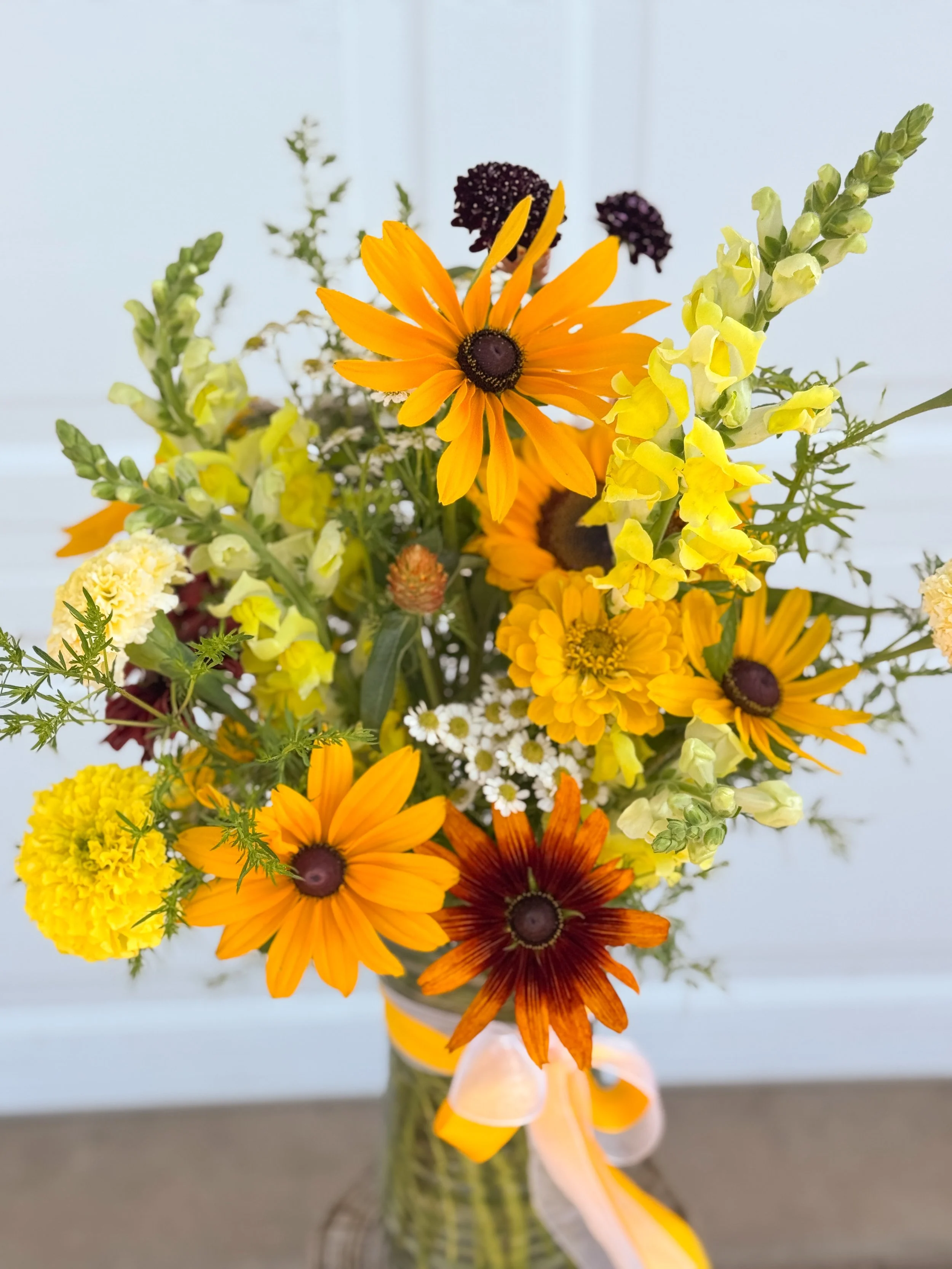 Vase with a colorful arrangement of yellow, orange, and purple flowers, including daisies and marigolds, with a yellow ribbon tied around the vase.