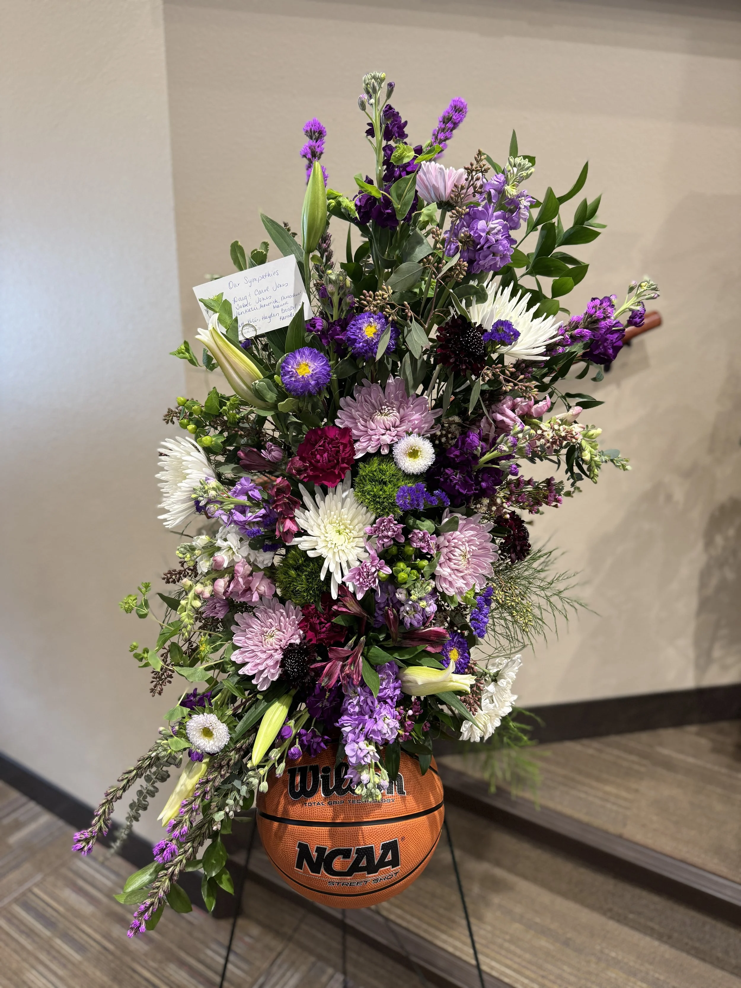 A floral arrangement in a pot shaped like a basketball with the Wilson and NCAA logos. The arrangement features pink, purple, white, and dark-colored flowers with green leaves, and has a handwritten note attached.