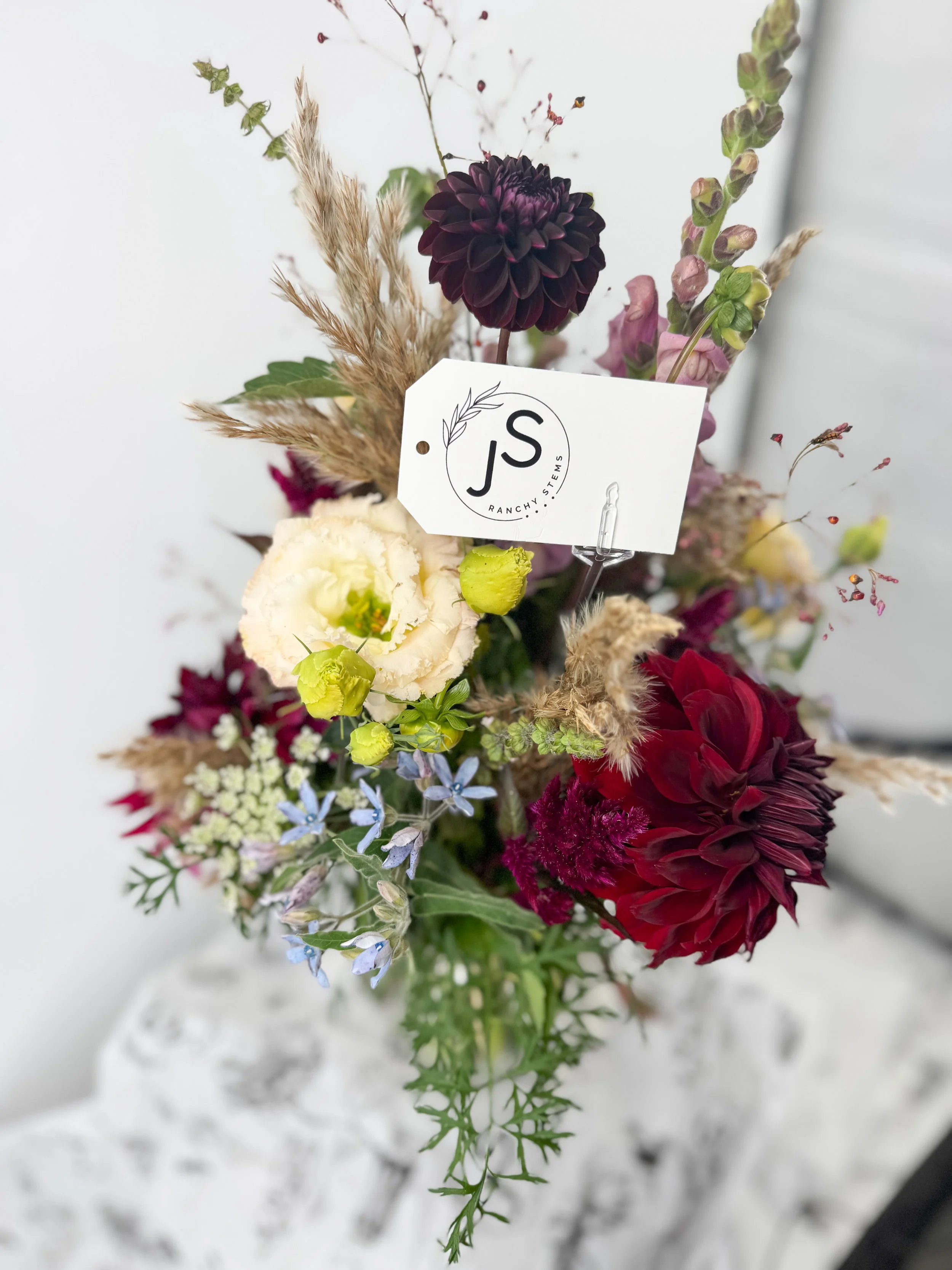 A mixed floral bouquet with dark purple, red, white, and light purple flowers, including ornamental grasses, in a vase on a white surface.