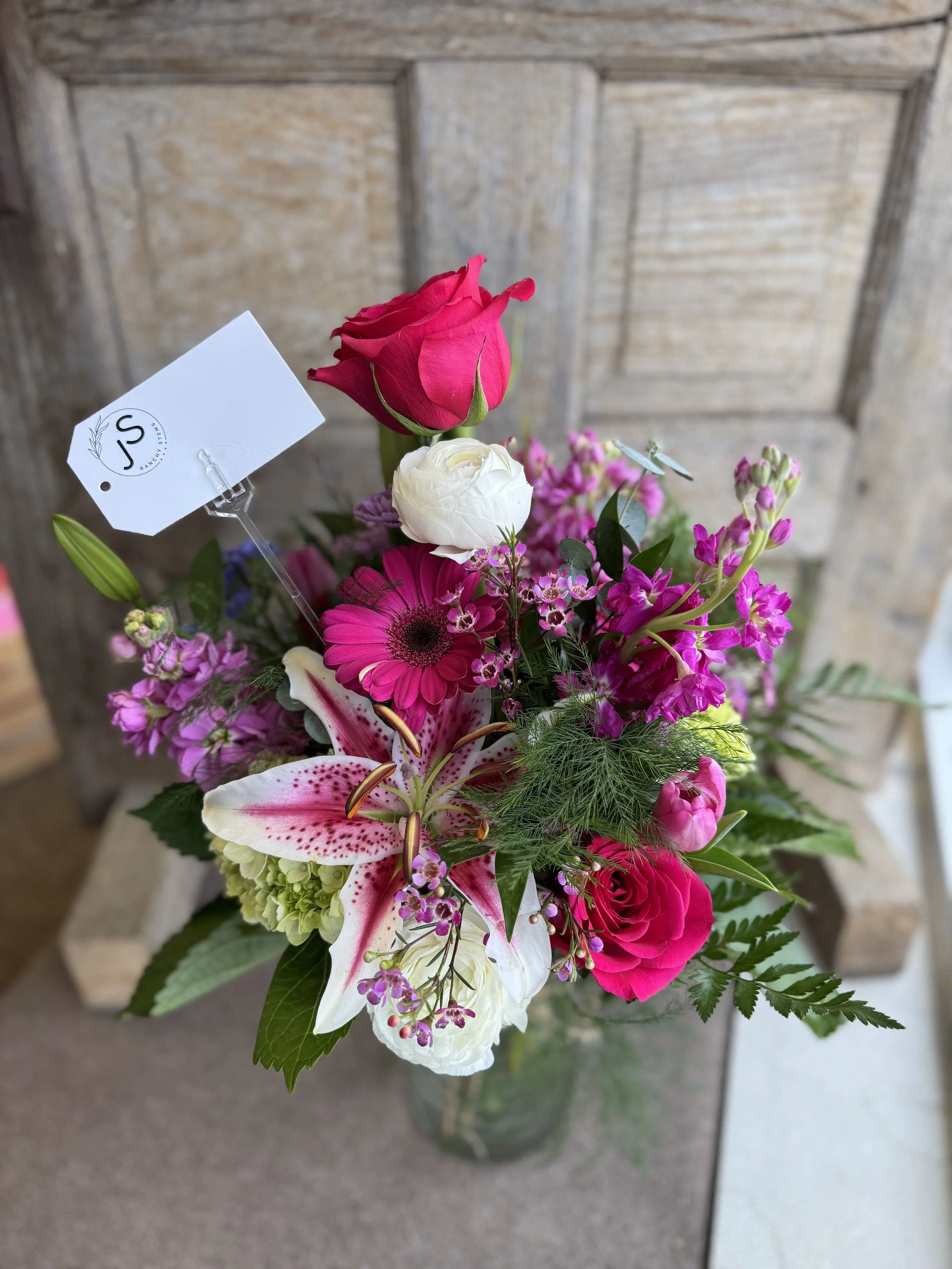 A colorful bouquet of pink, white, and purple flowers in a glass vase, with a small white tag attached to one of the flowers, against a wooden background.