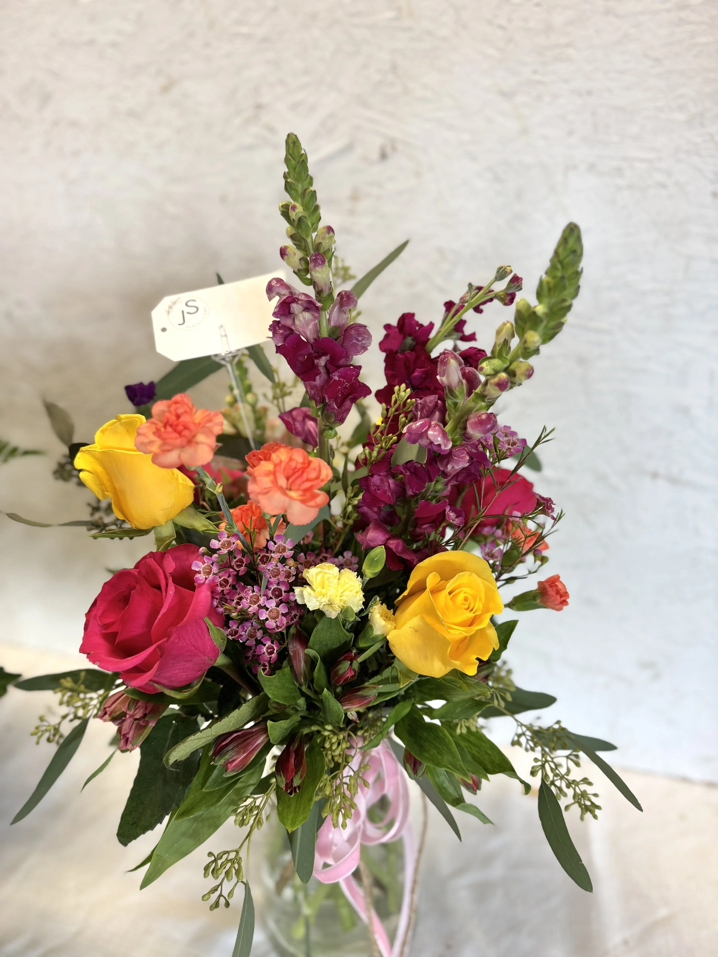 Colorful bouquet of yellow, pink, orange, and purple flowers in a glass vase with pink ribbon.