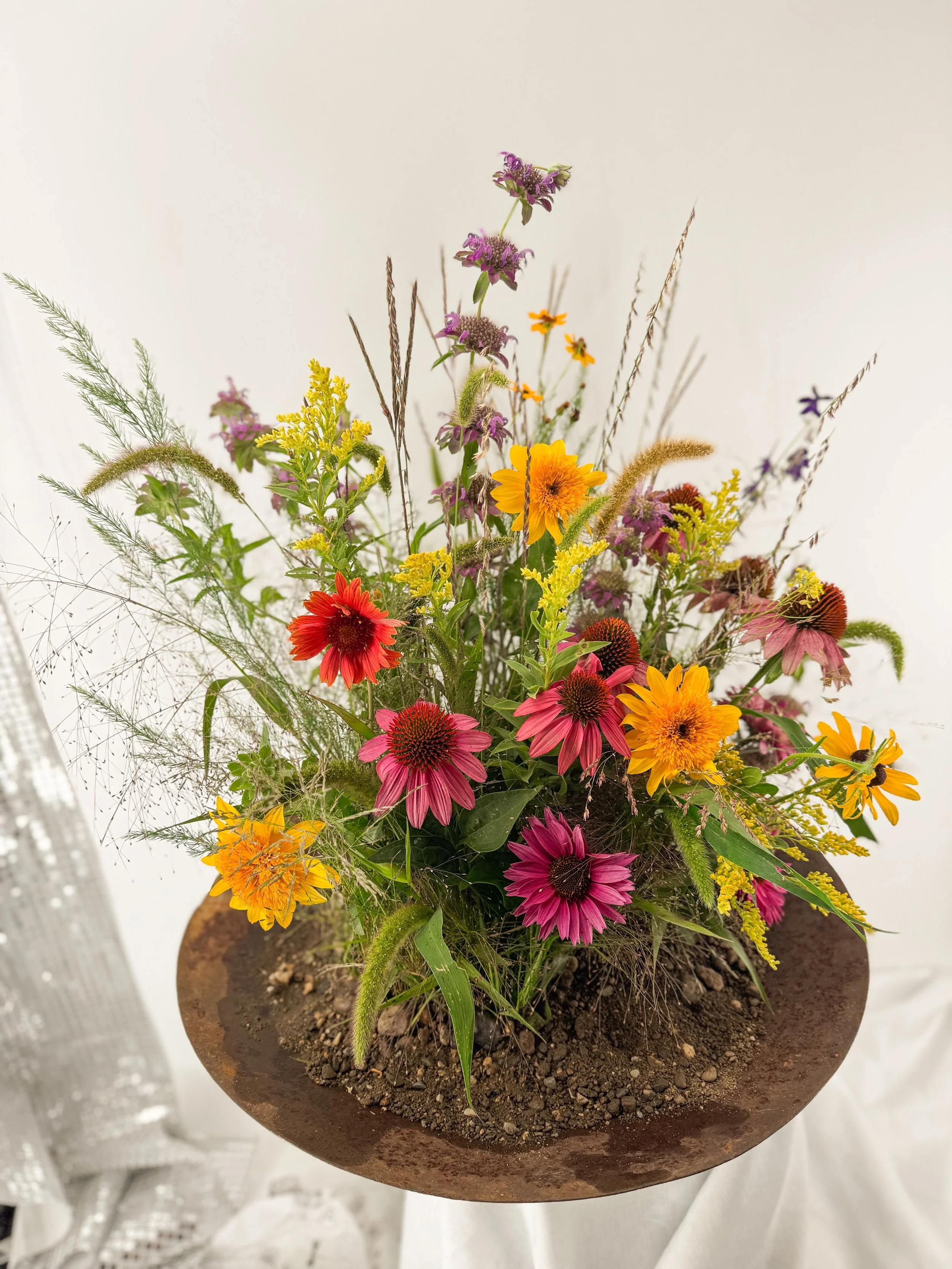 Colorful mixed flower arrangement in a round, rust-colored pot with soil on top, featuring yellow, pink, purple, and orange flowers and green foliage.