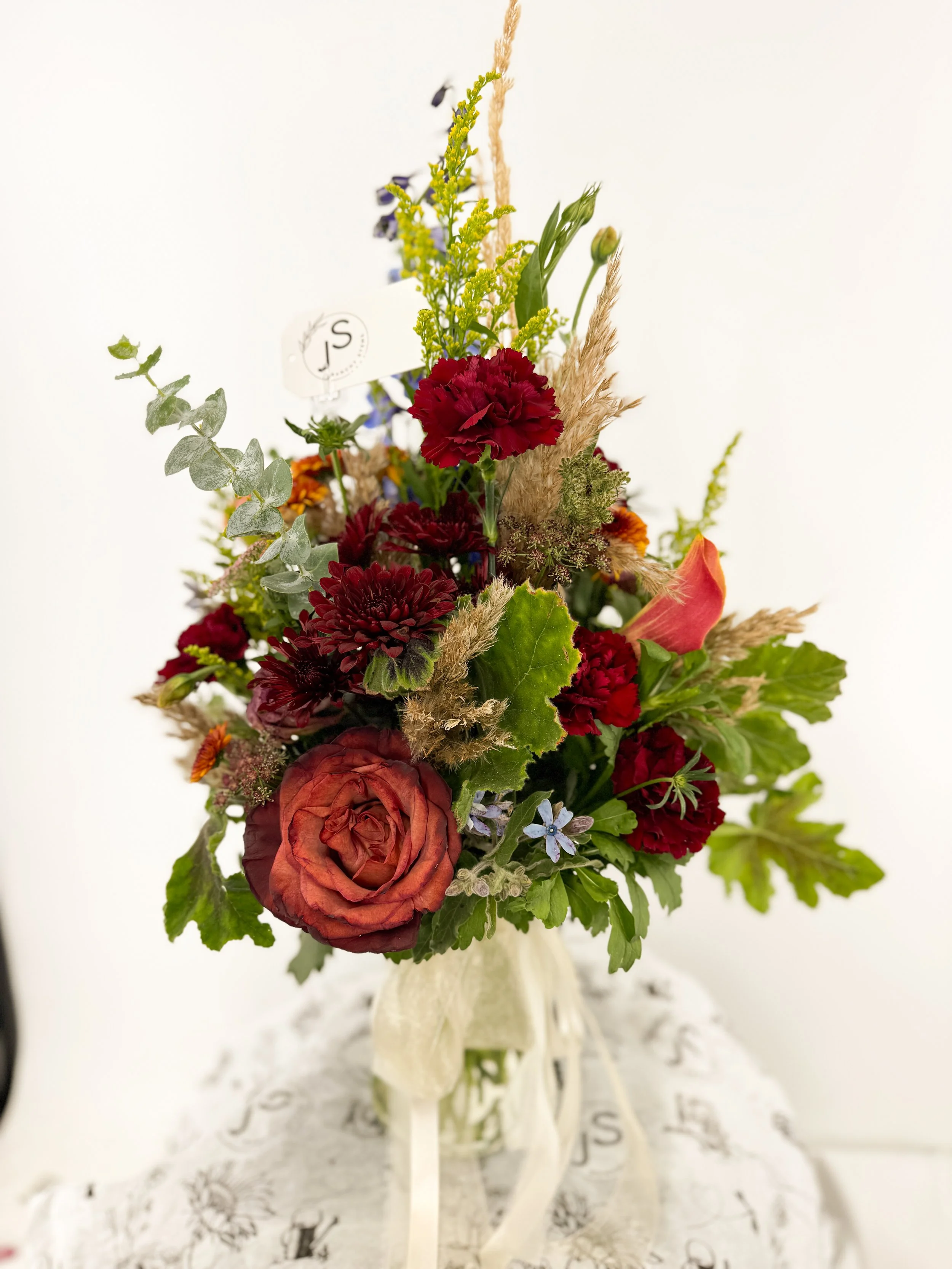 Colorful flower bouquet with roses, carnations, and greenery in a vase on a lace tablecloth.