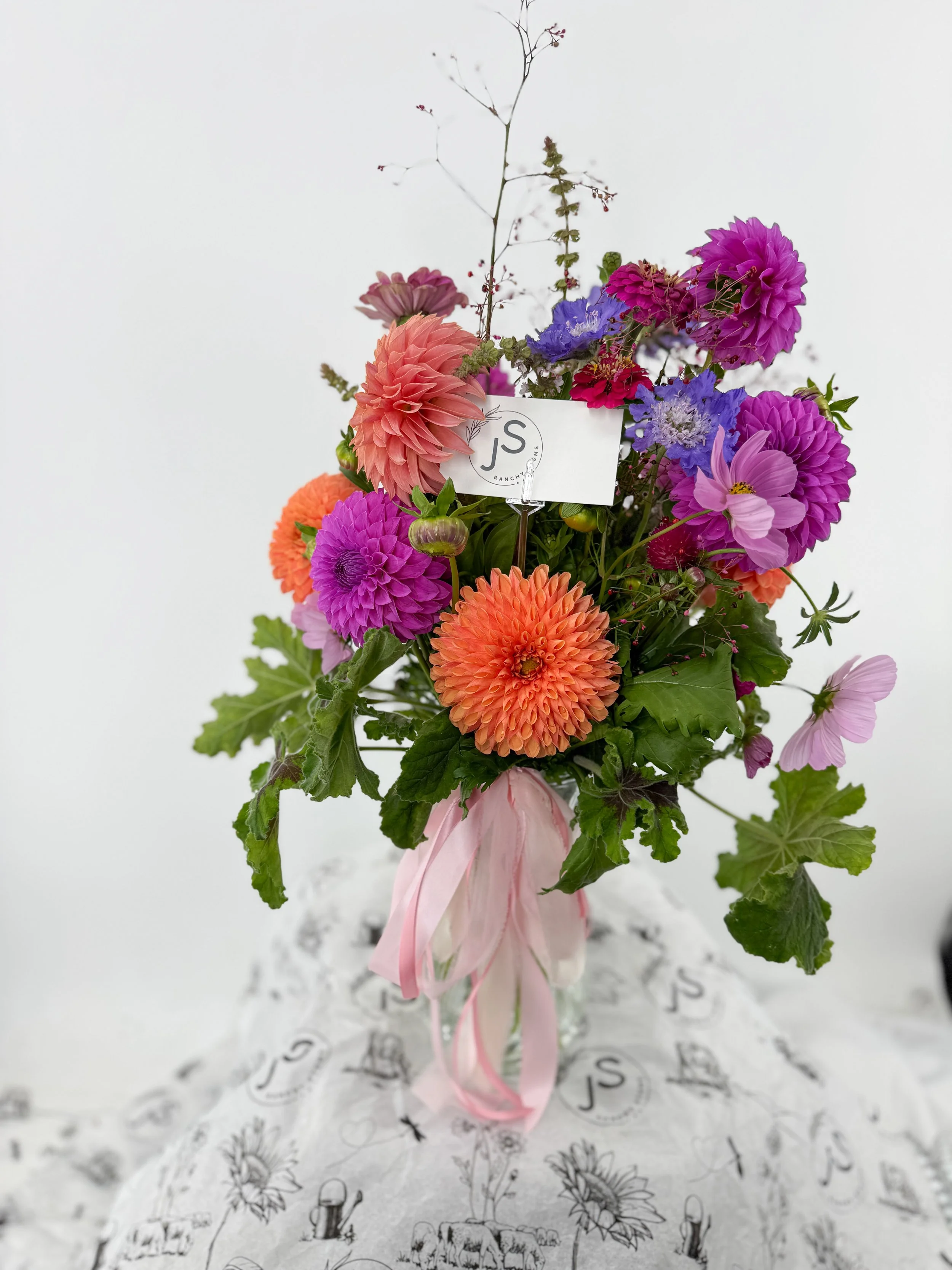 Colorful bouquet of flowers with pink, purple, orange, and blue blooms, tied with pink ribbons, placed on a patterned fabric with a white background.