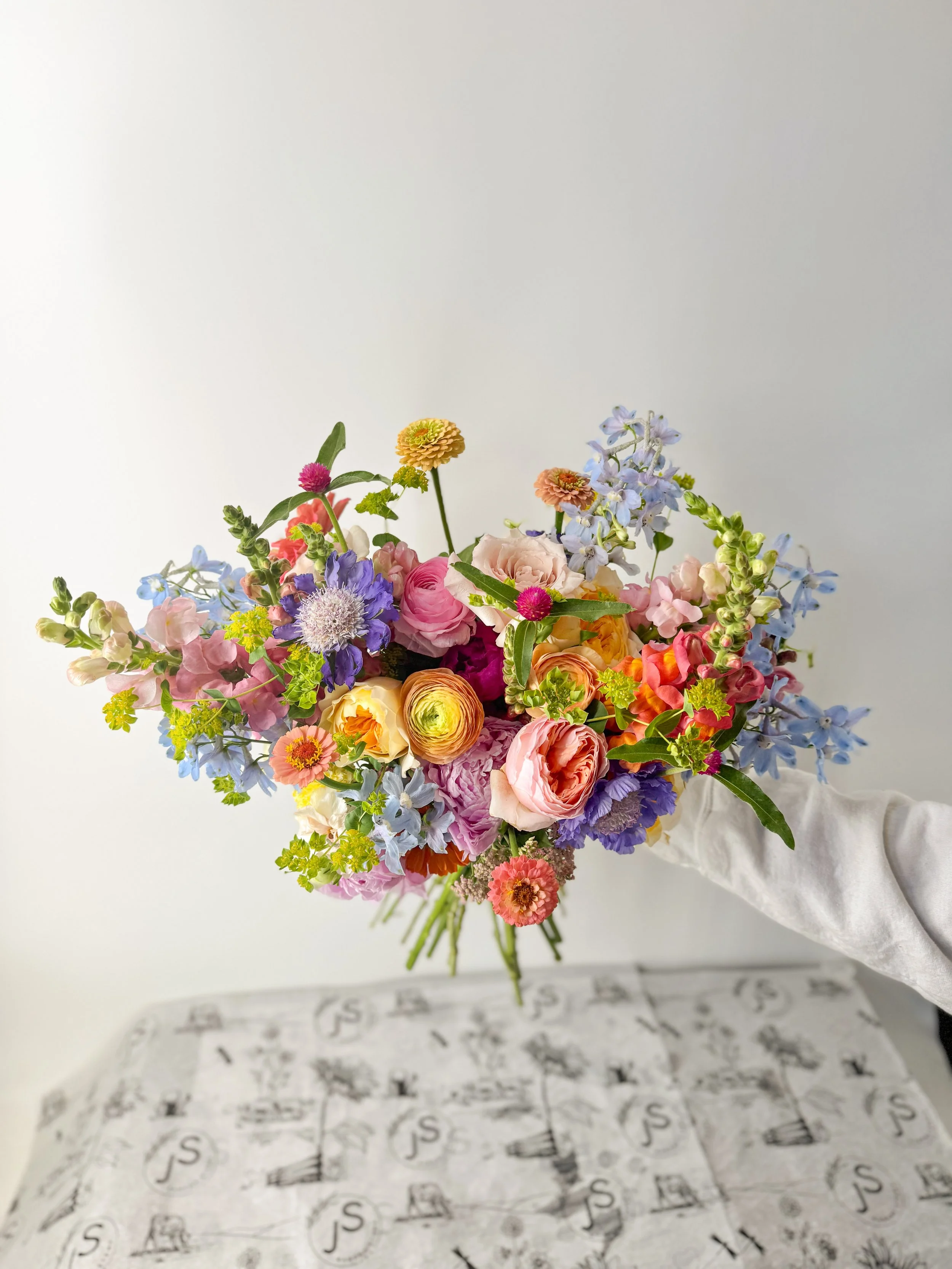 Colorful bouquet of various flowers including roses, ranunculus, and delphiniums, held by a person wearing a white sleeve, against a plain white background.